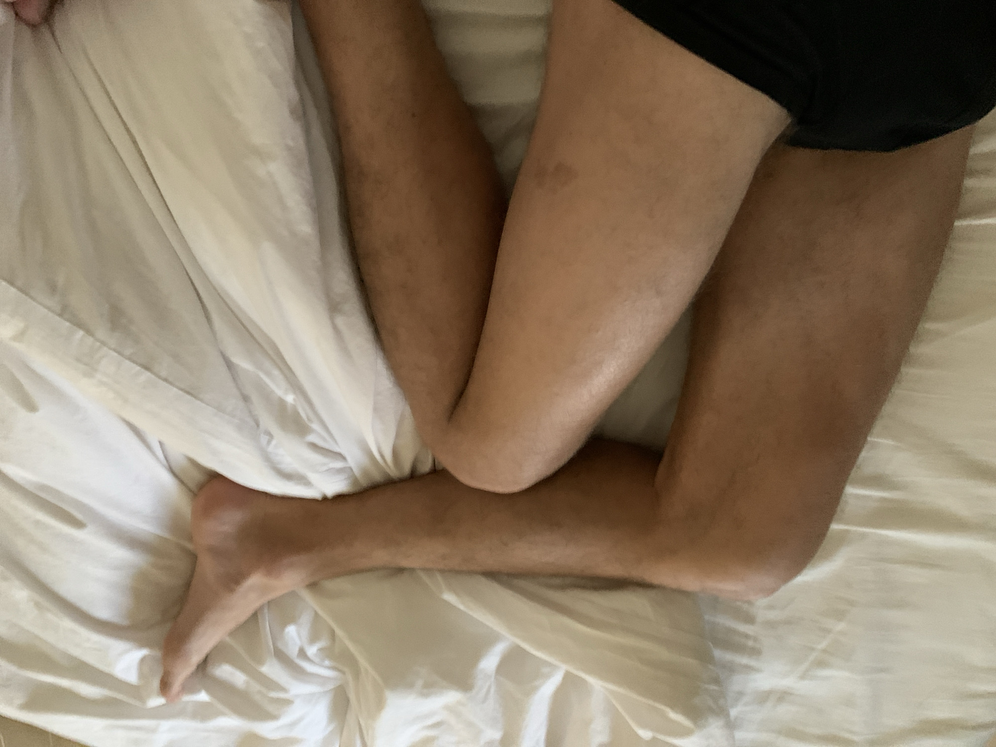 The image shows a person lying in a bed covered with white sheets. The person is wearing black underwear. Their legs are intertwined and their foot is visible on the left side of the frame. The location is not identifiable as Tel-Aviv, Israel from the visual information provided.