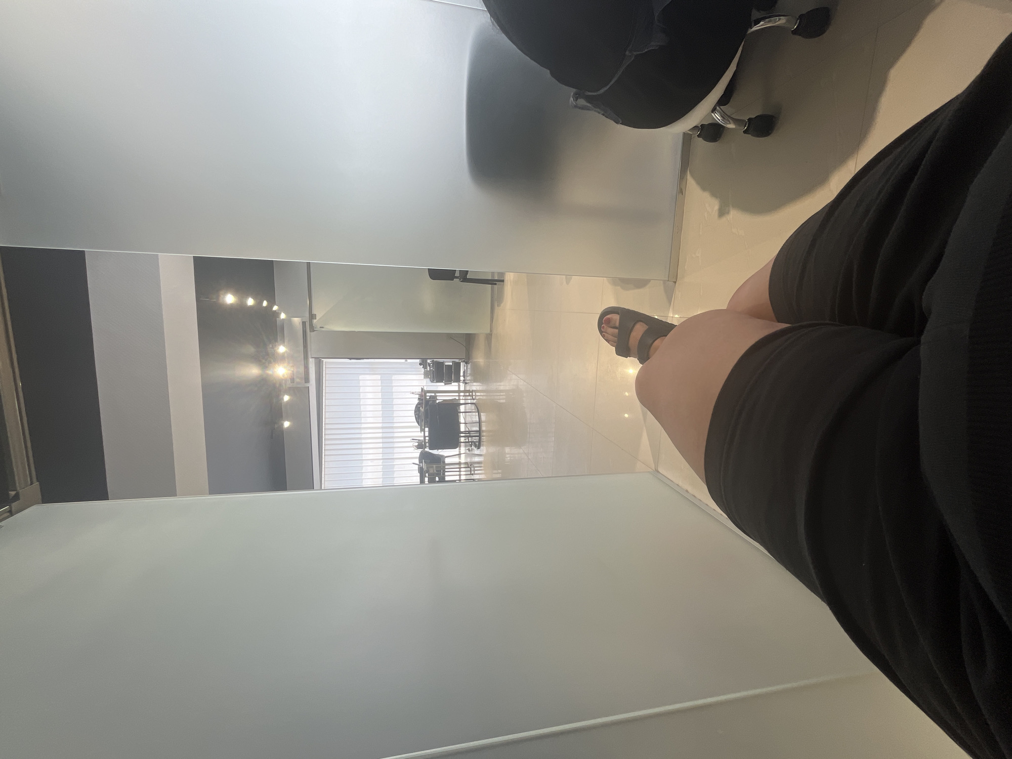 A first-person perspective shot shows a person's legs and feet, clad in black shorts and grey sandals, resting in what appears to be a waiting room or office. The floor is tiled and reflects light. Frosted glass partitions divide the space, revealing glimpses of a well-lit area with desks, chairs, and vertical blinds in the background. To the right, a black office chair with wheels is partially visible. The ceiling is dark with several spotlights.