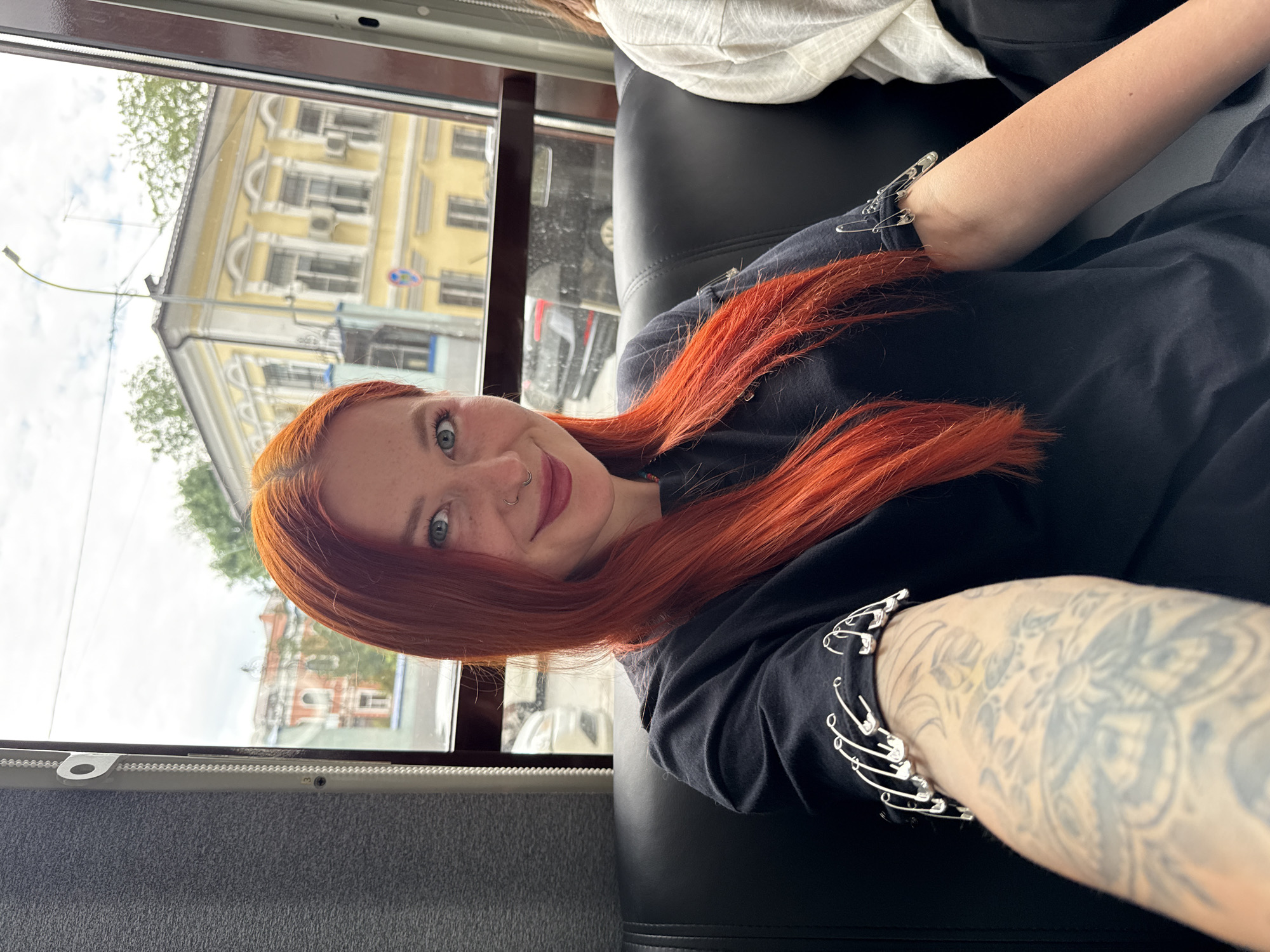 A young, fair-skinned woman with orange hair, a nose ring, and blue eyes is seen indoors, likely on public transportation. She is wearing a black long-sleeved shirt adorned with silver safety pins on the sleeves. The woman has tattoos on her arm. Outside the window, buildings typical of Rostov-on-Don, Russia are visible, including a multi-story yellow building with architectural details, and trees and other buildings further in the distance. Other passengers are also in the vehicle. The light is diffused, indicating an overcast day.