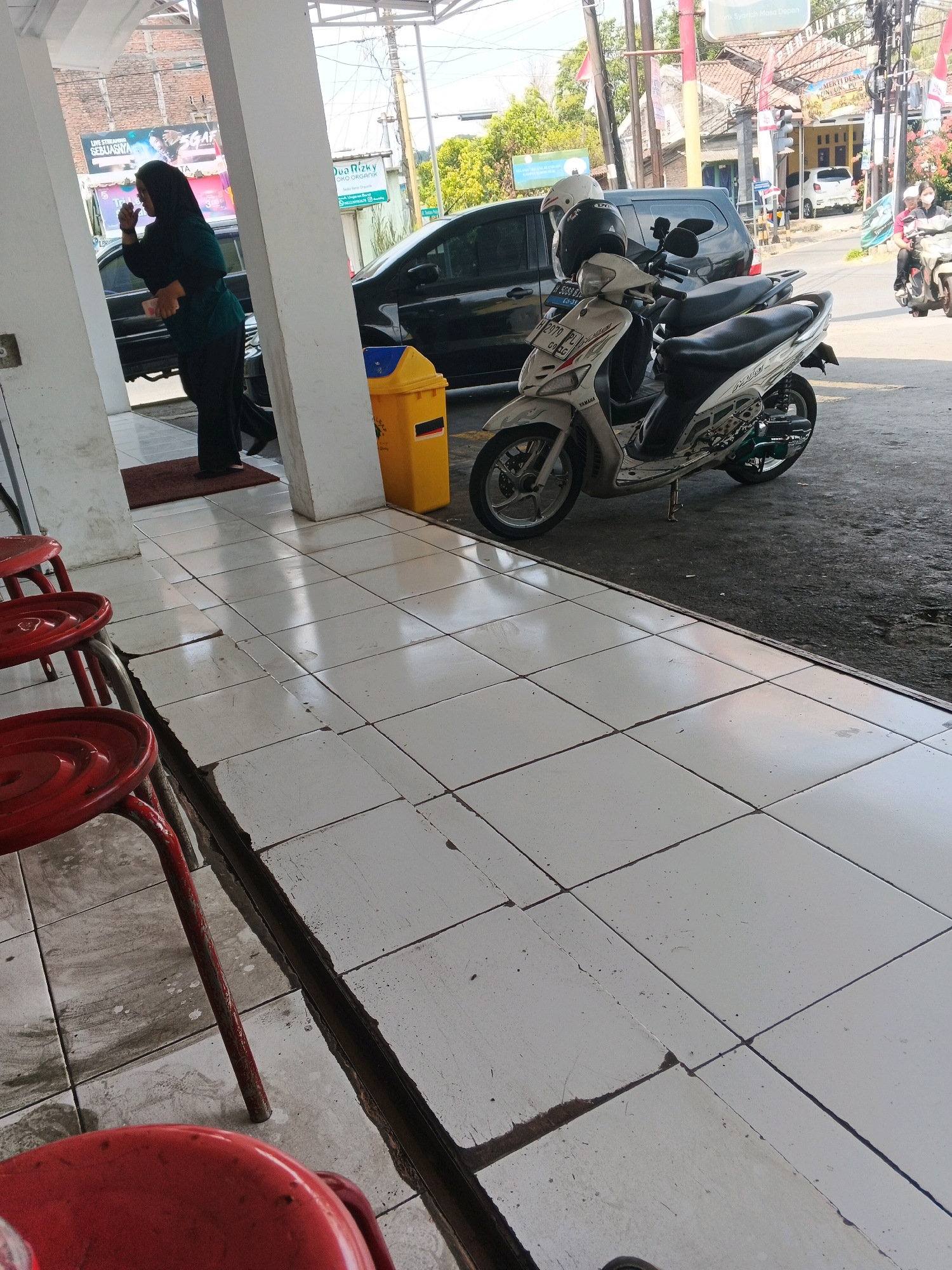 In Genuk Barat, Indonesia, a street scene unfolds with elements of daily life. A woman wearing a dark hijab and green top stands near a white pillar, facing away from the camera. Beside her is a black car, partially obscured. A yellow trash can is positioned nearby. In the foreground, three red stools are visible. Beyond the immediate area, motorcycles are parked. A white Yamaha scooter, with helmets on its handlebars, is prominent. Further back, another motorcycle carries two riders wearing helmets, one with a pink shirt. The environment appears to be a commercial or roadside area, with buildings and signage in the background. The ground is tiled, with some of the tiles cracked. The overall impression is of a typical Indonesian street environment.