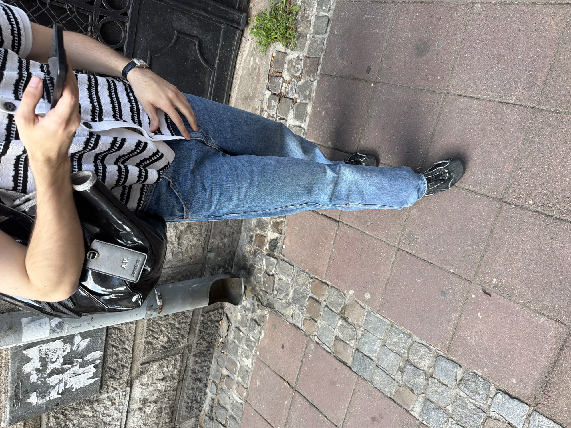 A person is standing on a street paved with red rectangular bricks and gray cobblestone in Belgrade, Serbia. The person is wearing blue jeans, a white and black striped button-down shirt, and dark-colored shoes. They are holding a black smartphone in their left hand. A black patent leather bag with a gray luggage tag marked "AV. Samsonite" hangs from their left shoulder. The person is standing near a dark metal gate and a gray metal pipe against a stone wall with peeling posters.