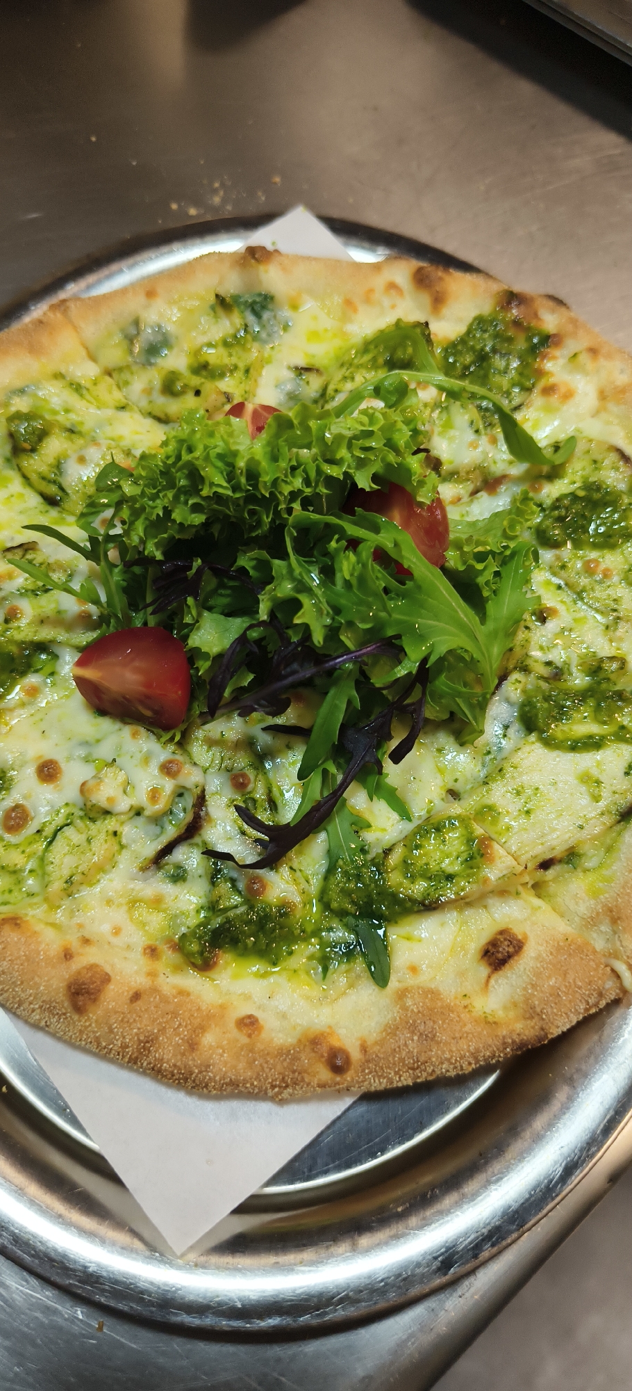 A close-up, high-angle shot captures a freshly prepared pizza presented on a round, reflective metal tray. The pizza features a thick, golden-brown crust and a base of melted white cheese, generously interspersed with a vibrant green pesto-like sauce. Piled in the center is a fresh salad mix, comprising bright green curly lettuce, darker leafy greens (likely arugula or similar), and two halves of red cherry tomatoes. A white, rectangular piece of paper is visible beneath the pizza, separating it from the metal tray. The tray rests on a blurred background that appears to be a metallic or light-colored counter surface, suggesting a commercial kitchen or restaurant environment. The image depicts the pizza ready for consumption, characteristic of a mealtime setting in Yerevan, Armenia. No people or visible text are present in the frame.