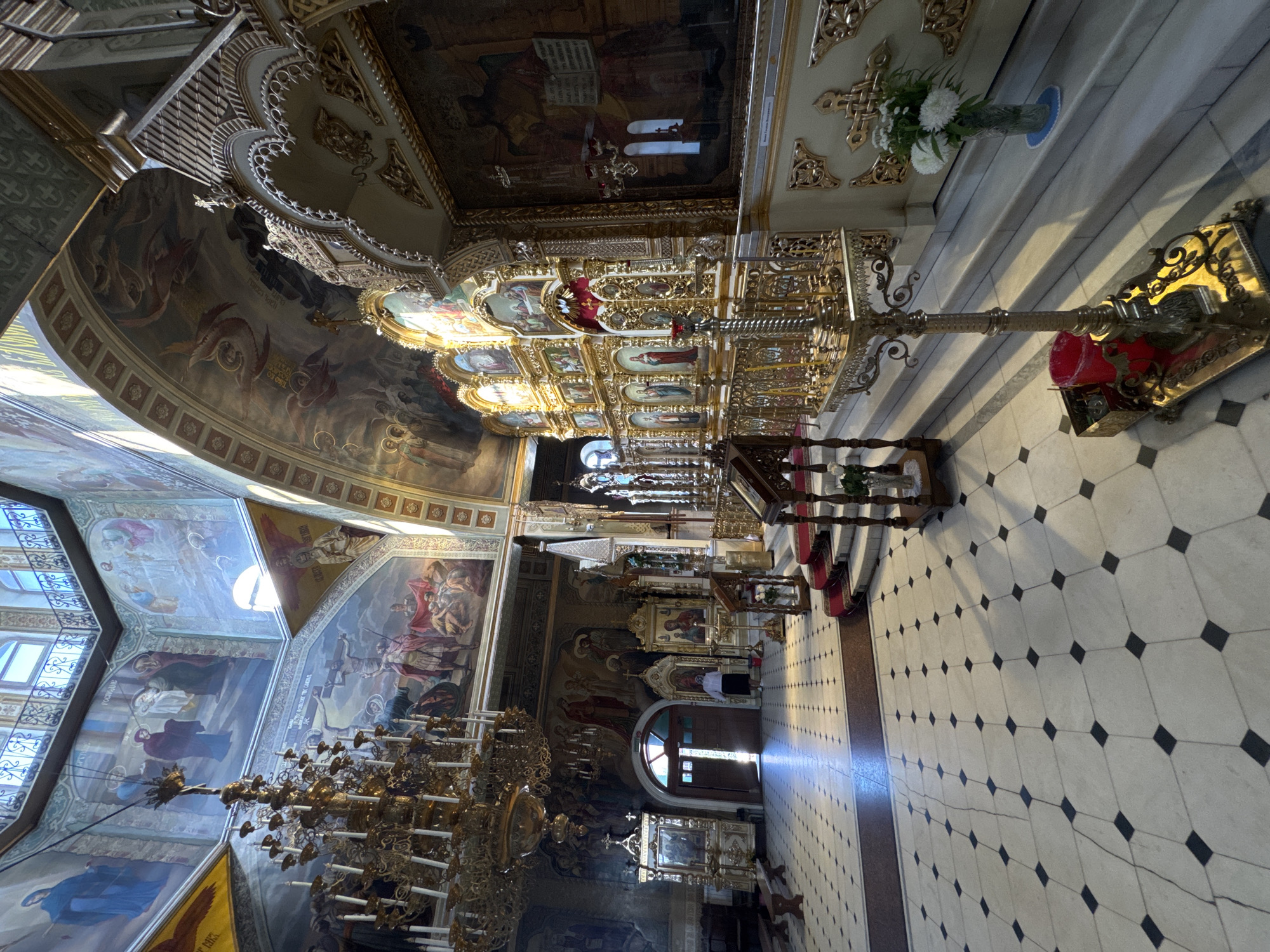 The image depicts the interior of the Transfiguration Cathedral in Tolyatti, Russia. The cathedral features a grand chandelier, elaborate gold-gilded iconostasis, and ornate frescoes adorning the walls and ceiling. The floor is tiled with a checkered pattern of white and black squares. A woman in dark clothing and a headscarf is standing near an open doorway in the background. The scene is illuminated by sunlight streaming through windows and the chandelier.