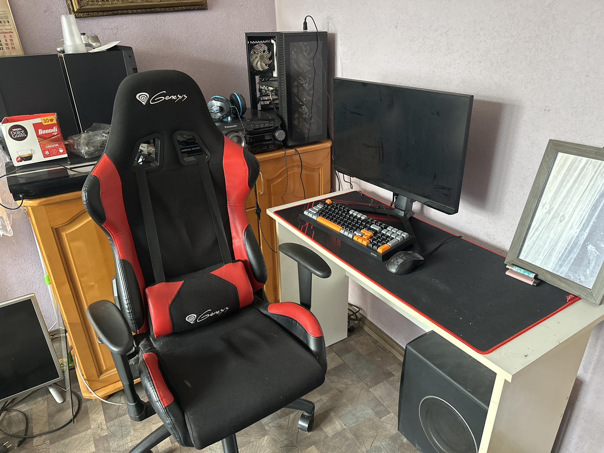 The image depicts a gaming setup in a room, likely a home office, in Cherven Breg, Bulgaria. The central focus is a black and red gaming chair, labeled with the "Genesis" logo. To the right of the chair, a desk holds a computer monitor, a keyboard with orange and grey keys, and a mouse. Behind the desk, a black computer tower is visible. On the left side of the image, a wooden cabinet contains various electronic devices, including what appears to be a game console and boxes of coffee pods. A framed mirror is positioned on the right side of the desk, reflecting part of the room. The room's walls have a textured, light purple-pink color. The flooring is a wood-patterned laminate. The overall scene is static, showing a pre-arranged setup without any visible activity.