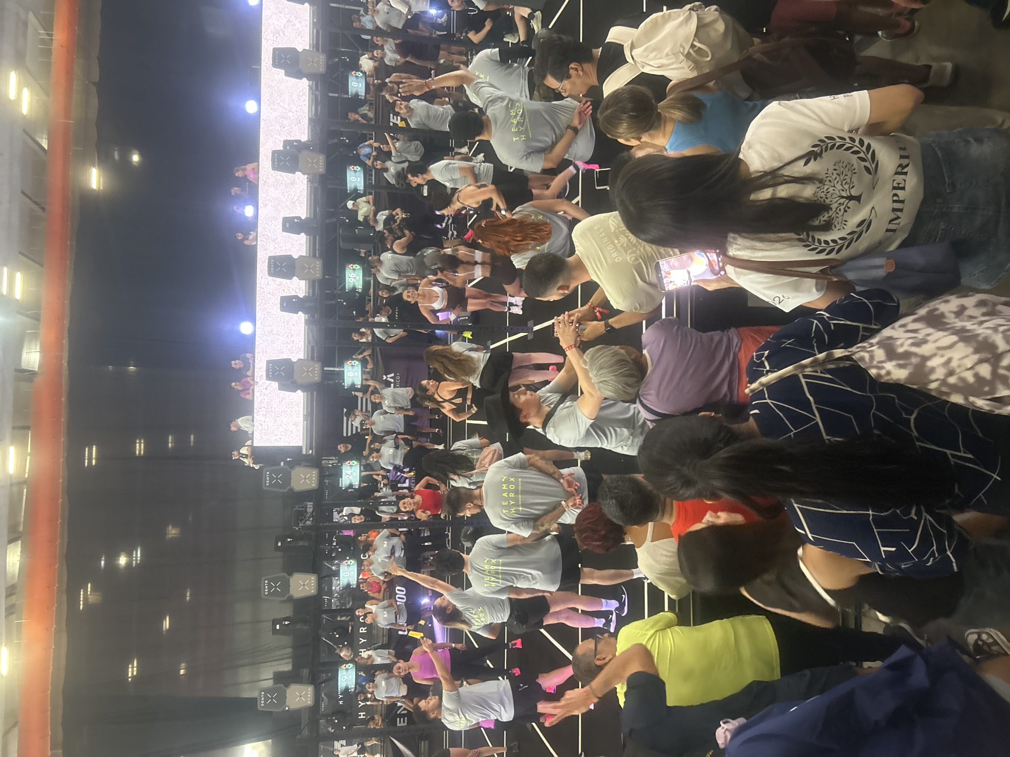 A densely packed fitness event is underway indoors, as observed from an elevated perspective. The event features numerous participants actively engaged in a fitness challenge, likely a HYROX competition, as evidenced by the "HYROX" signage and logos visible on the equipment and attire of some participants.
Participants are distributed throughout the space, grouped around fitness stations that appear to integrate digital displays. Many individuals are clad in athletic wear, and some are wearing shirts with the "TEAM HYROX" insignia, suggesting they may be part of organized teams. Various fitness actions seem to be taking place, with individuals stretching, lifting weights, and engaging in other physical exertions.
The venue appears to be a large gymnasium or warehouse-type structure, based on the high ceilings and expansive open space. Lighting is provided by overhead fixtures, and a stage with illuminated panels is visible in the distance, possibly indicating a performance or awards area. The presence of individuals observing the activities suggests a spectator element to the event. Some people are using mobile phones.