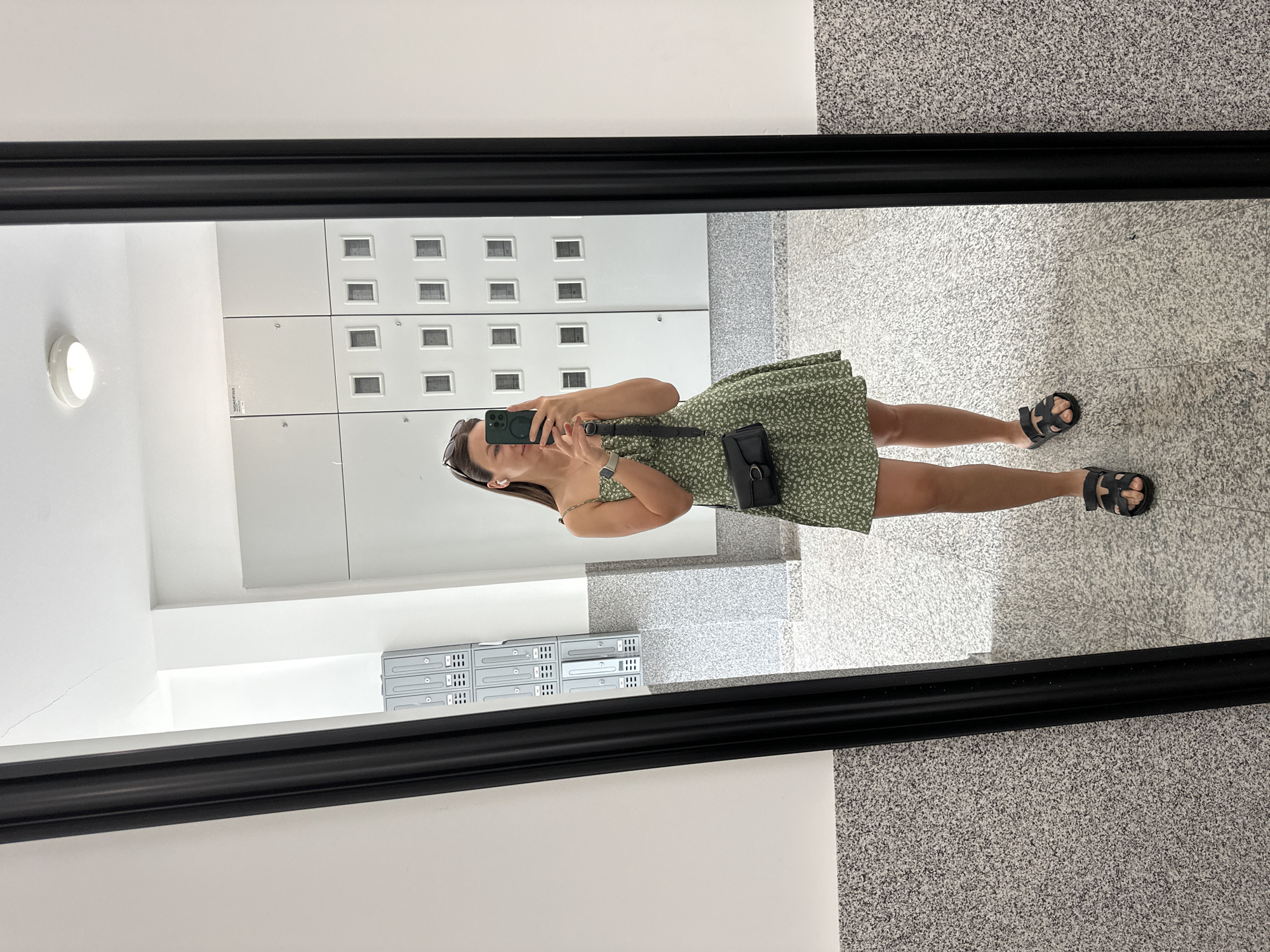 A woman in a short, green floral dress and black sandals takes a mirror selfie in a hallway. The hallway has gray speckled floors, white lockers lining one wall, and a white wall with mounted electronic equipment near the base of the lockers. The mirror has a black frame. The location is not discernable as Lapčići, Montenegro based on visual information.