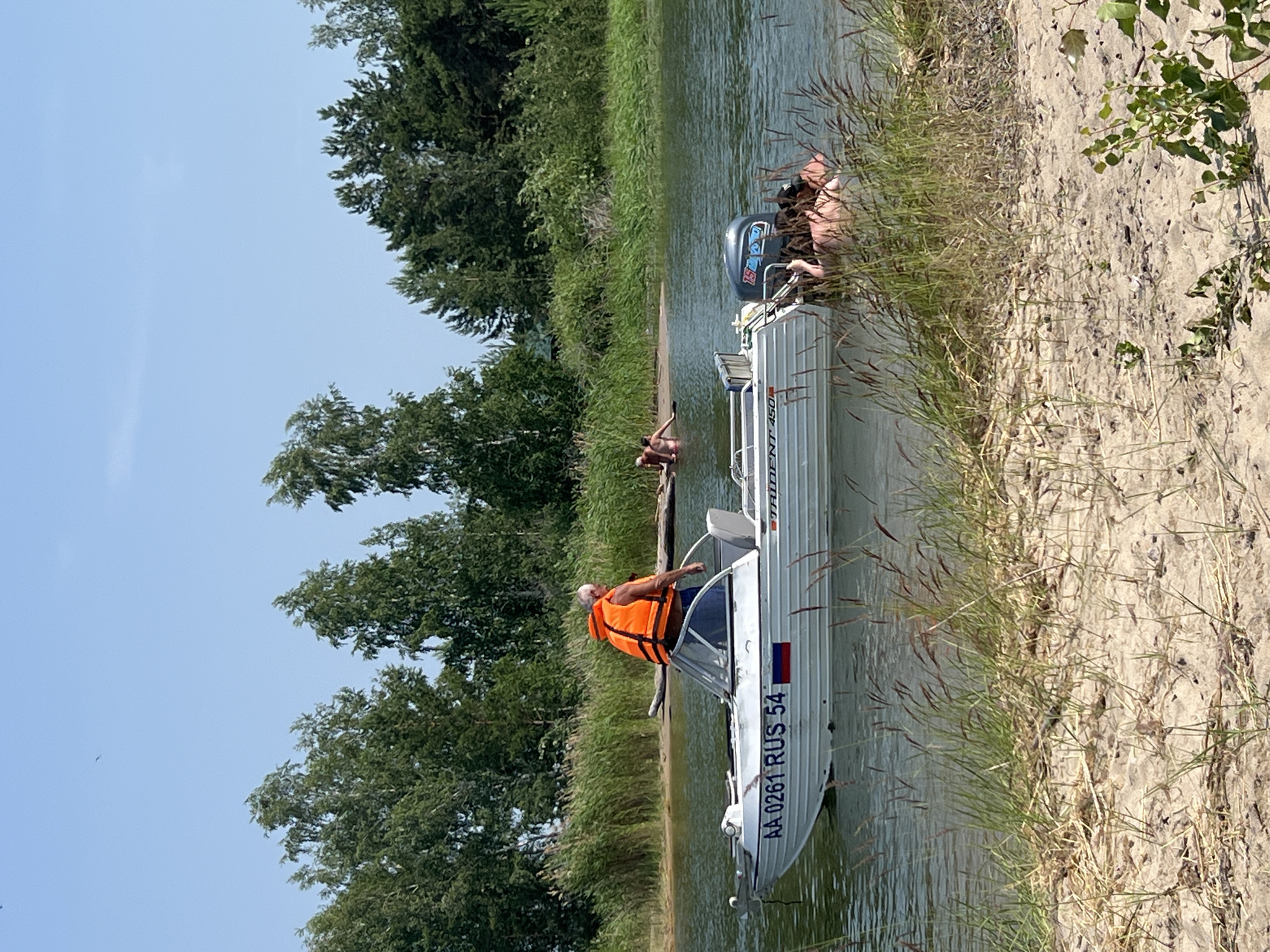 The image shows a scene on a body of water, likely a lake, on a sunny day. A group of people are enjoying a summer outing.
In the foreground, a white motorboat is partially submerged near a sandy shore covered with sparse vegetation. An older man wearing an orange life vest is standing in the boat, looking towards the water. The boat has visible text on its side: "AA 0261 RUS 54" and "TAIDER". Another person is partially visible in the boat, wearing blue shorts.
Further in the water, another person, also appearing to be in shallow water, is partially submerged. In the background, lush green trees and reeds line the shore. The sky is a clear, pale blue.
The scene appears to be a recreational activity, likely swimming or boating, taking place in a natural environment. The overall mood seems relaxed and leisurely, consistent with a summer day spent outdoors. The location cues, such as the text on the boat indicating Russian registration and the name "Borovoe" (though not directly visible in the image, it's provided as context), suggest the setting is in Russia. The bright sunlight and clear sky indicate fair weather.