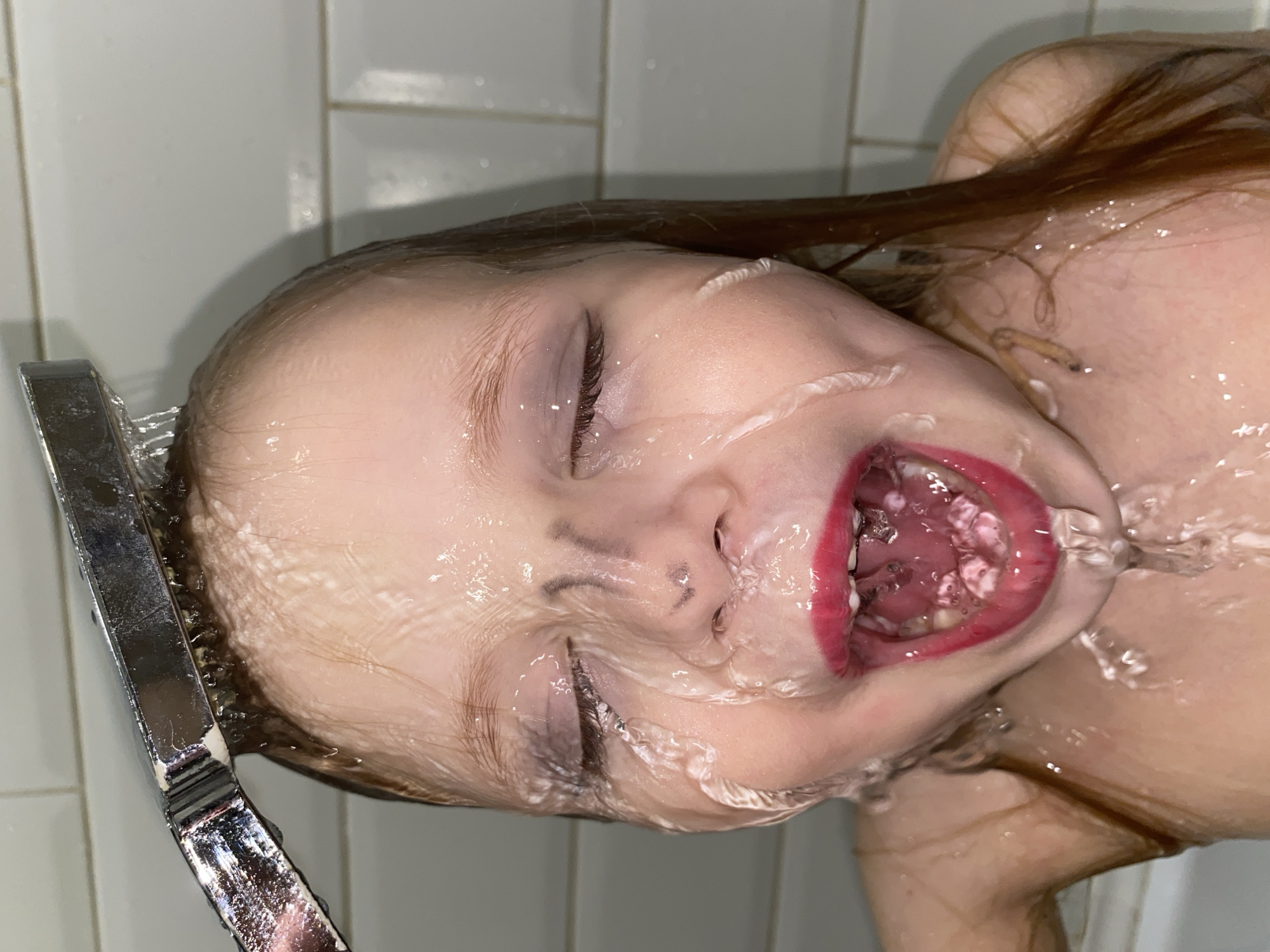 A young person with brown hair is in a shower with water running over their face. Their eyes are closed, and their mouth is open. The shower head is visible on the left side of the image. The background consists of white tiles. There is no information to suggest this image was taken in Votkinsk, Russia.