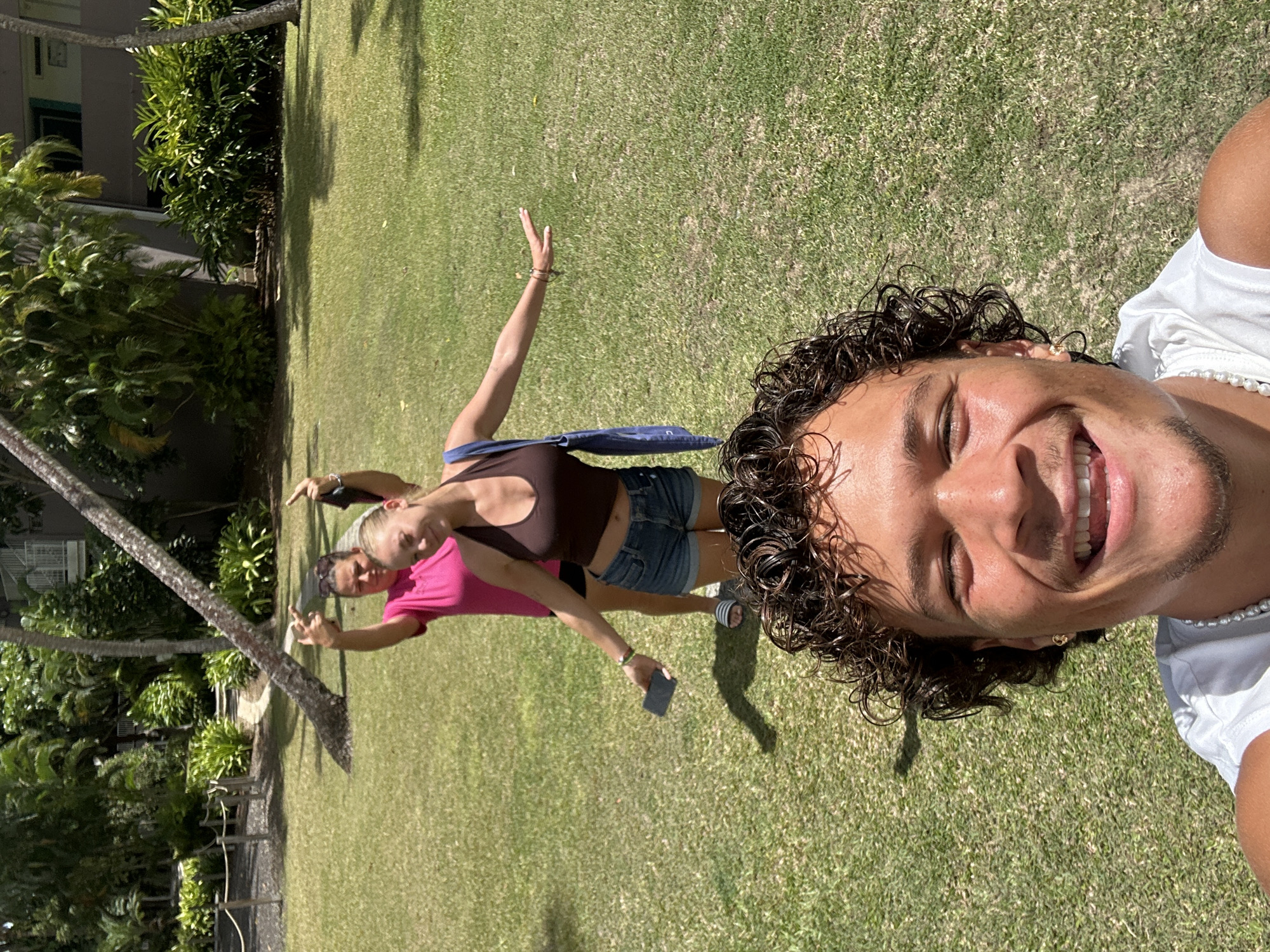 The image depicts three individuals on a grassy area. The person in the foreground is a young male with curly brown hair, wearing a white shirt and a pearl necklace. He is smiling at the camera. Behind him are two females, one wearing a brown top and denim shorts, and the other a pink shirt. They both have their arms raised. The woman in the brown top is holding a dark-colored mobile phone. The background includes trees and a building. There is no evidence to indicate the location is in the city of Sainte-Anne, France.