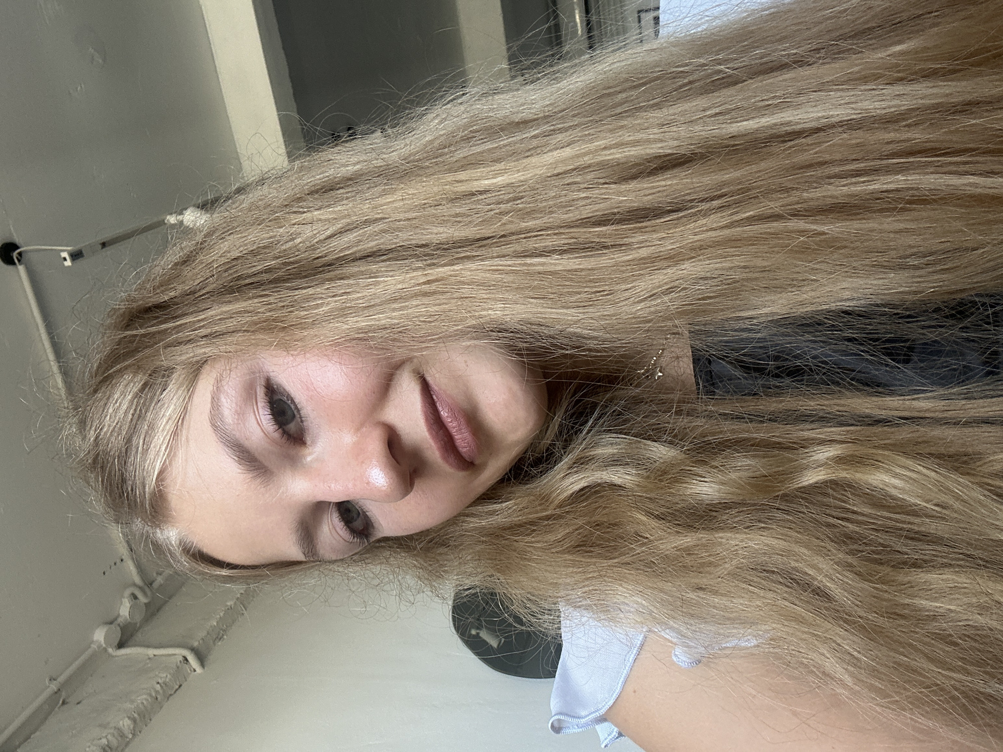 A blonde-haired woman is pictured in a close-up, downward-tilted selfie, indoors. The setting appears to be a room with white-painted walls and a partially visible metal structure. The woman's long, blonde hair is the focal point, cascading across the frame. The location is not discernible from the provided details to confirm Kuta Selatan.