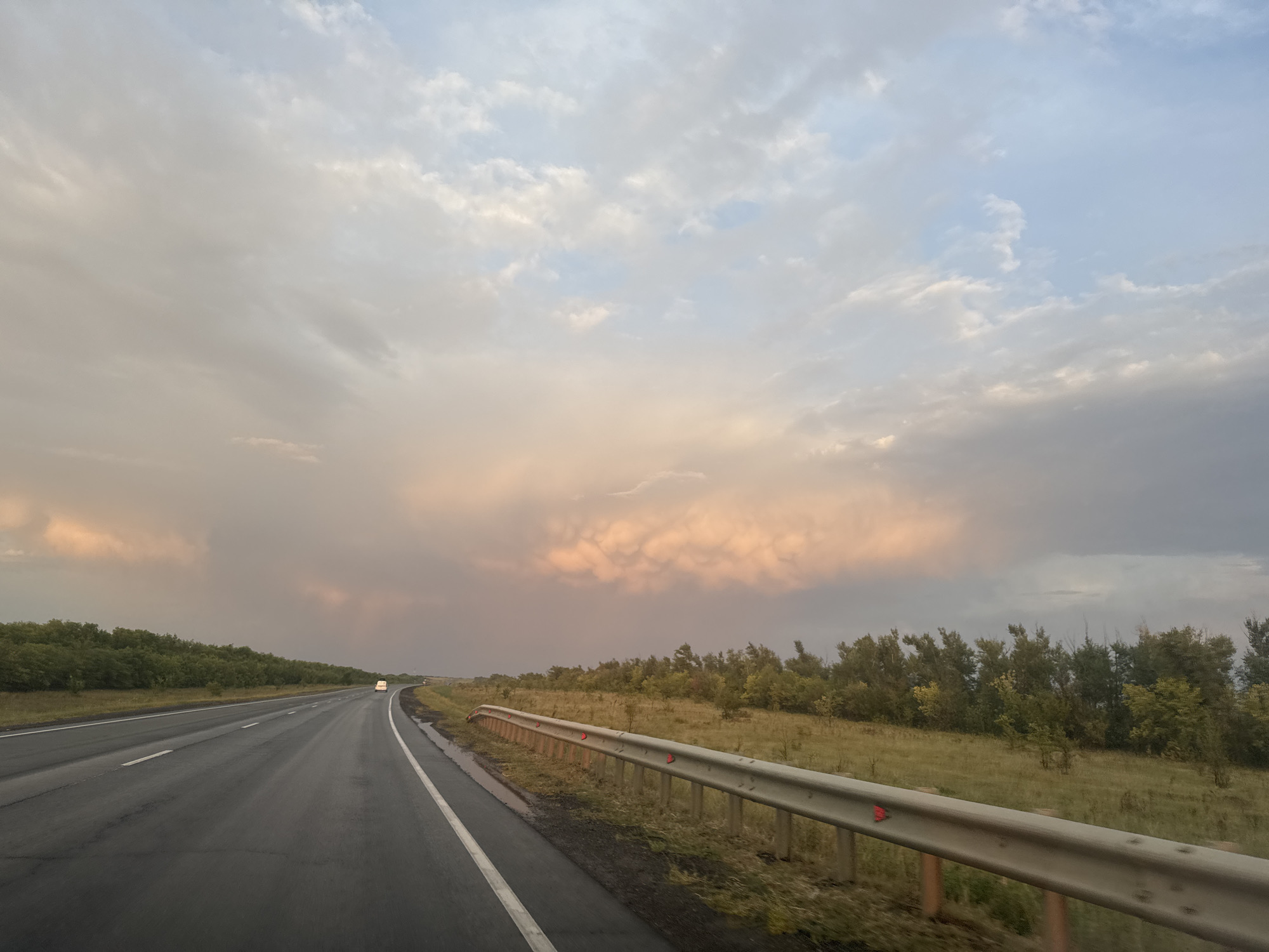 An asphalt highway stretches into the distance on a cloudy day. On the left side of the road, a line of trees forms a dense green barrier. On the right side of the road, a grassy field is bordered by trees and a metal guardrail. A white van is visible on the highway in the distance, and the sky is filled with dramatic clouds tinged with pink and orange hues, suggesting either sunrise or sunset. The scene appears to be on a rural road outside of Perevolotsky, Russia.