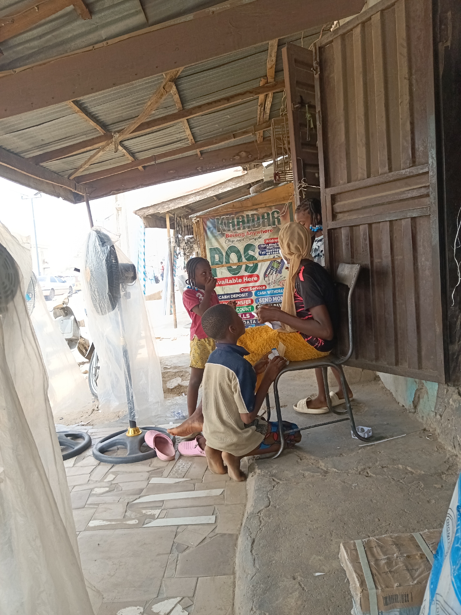 The image shows an outdoor scene in Minna, Nigeria, where a woman sits on a metal chair in front of a shop. She is wearing a head covering, a dark t-shirt, and a yellow skirt. Two children stand near her; a girl in a red shirt and yellow shorts, and a boy in a blue and beige shirt who is kneeling. The shop's sign reads "KHAIDAR POS," advertising services such as cash deposits and withdrawals. A standing fan, partially covered in plastic, is visible to the left. The flooring is concrete. The area appears to be a small business or storefront. Additional objects present include a box on the floor and pairs of sandals.