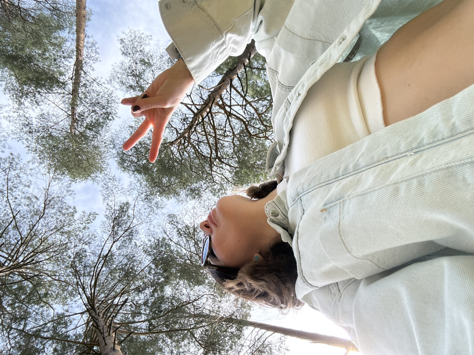 An upward-facing, low-angle photograph captures a woman against a backdrop of tall pine trees and a bright sky. The woman is positioned on the right side of the frame, her body partially visible from the chest up, looking directly upwards with a serene expression. She is wearing a light-wash denim jacket over a white top. Her short, dark hair is styled around her face, and sunglasses are perched on top of her head. A small, round turquoise earring is visible on her right earlobe. Her right hand is raised, forming a V-shape or peace sign with her index and middle fingers, which are painted with dark nail polish.
The background is dominated by the majestic, slender trunks and green, needle-like foliage of numerous pine trees that stretch high towards the sky. The branches of these trees create intricate, almost skeletal patterns against the bright, clear blue sky, which shows only a few wispy clouds. The lighting suggests it is daytime, likely late morning or early afternoon, with a gentle, ambient glow. A soft sun glare is faintly visible at the bottom right corner of the image. The scene evokes a sense of tranquil immersion in a natural, forested environment, characteristic of regions like Chaikovsky, Russia. There is no visible text in the image.