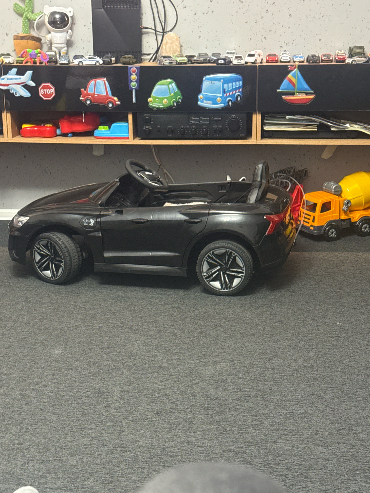 The image depicts a black ride-on electric car for children, situated indoors on a gray carpeted floor. The car is a scale replica of a modern vehicle, featuring detailed wheels, a steering wheel, and interior seating. Adjacent to the ride-on car, a yellow toy cement mixer truck is visible. The background reveals a shelf unit displaying various miniature toy vehicles, including a red toy car, a green toy car, an airplane, a stop sign, a blue bus, and a sailboat. Above the shelf, a collection of small toy cars are lined up, and a decorative cactus and an astronaut figure are also present. The overall scene suggests a child's play area or a collection of toys displayed in a home environment. There is no indication of the city of Gorzow, Poland, or any specific urban context.