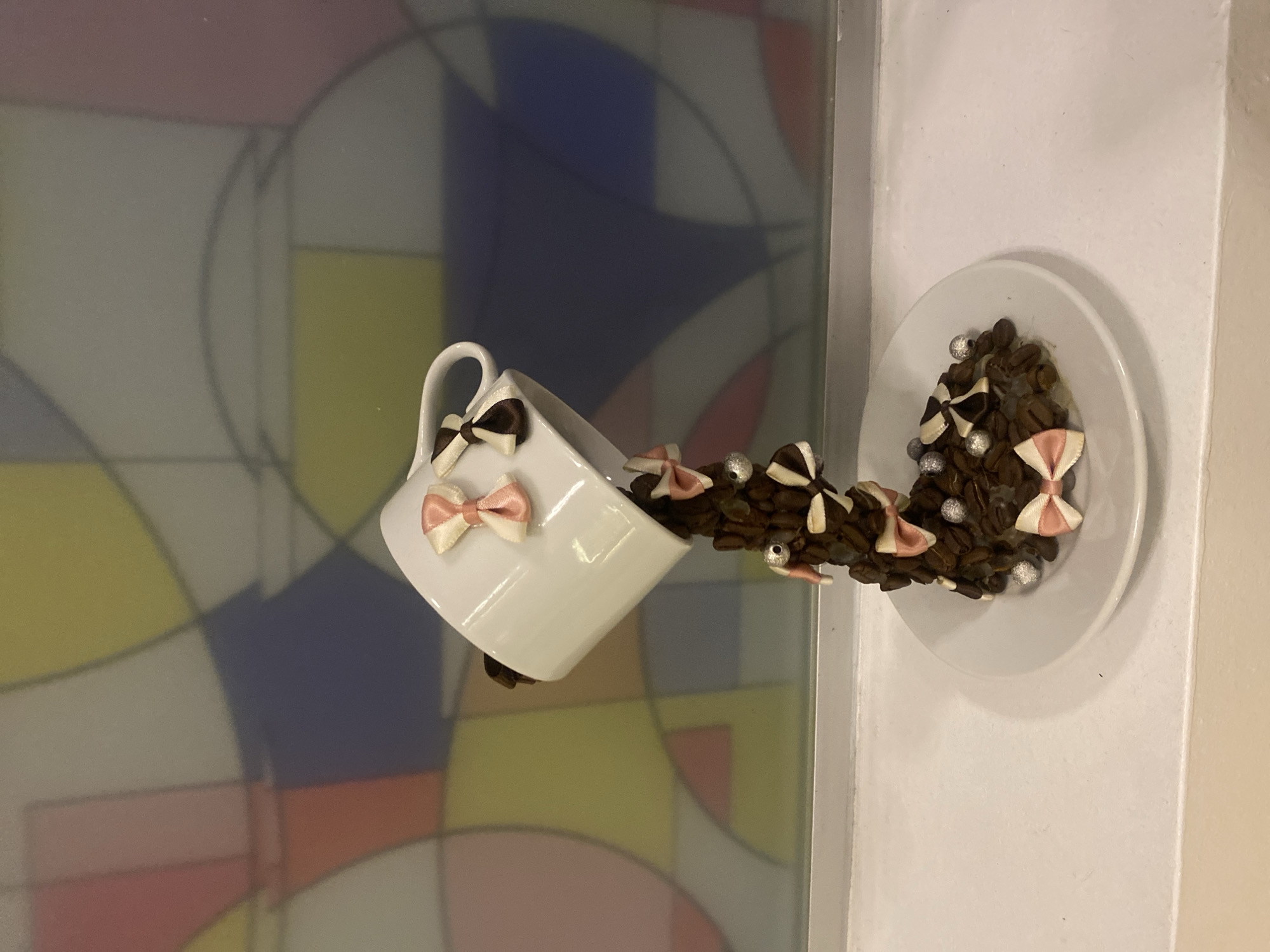 The image depicts a creative display of coffee beans cascading from an overturned white mug. The mug, adorned with small pink and white bows, is tilted as if pouring its contents. The coffee beans spill out and form a stream, also decorated with bows and small silver beads, flowing onto a white saucer. The background features a colorful, abstract stained-glass pattern with geometric shapes in shades of blue, yellow, pink, and white. The display rests on a white ledge, likely a windowsill. This scene is not located in Kuala Lumpur, Malaysia, as no elements in the image suggest this specific location.