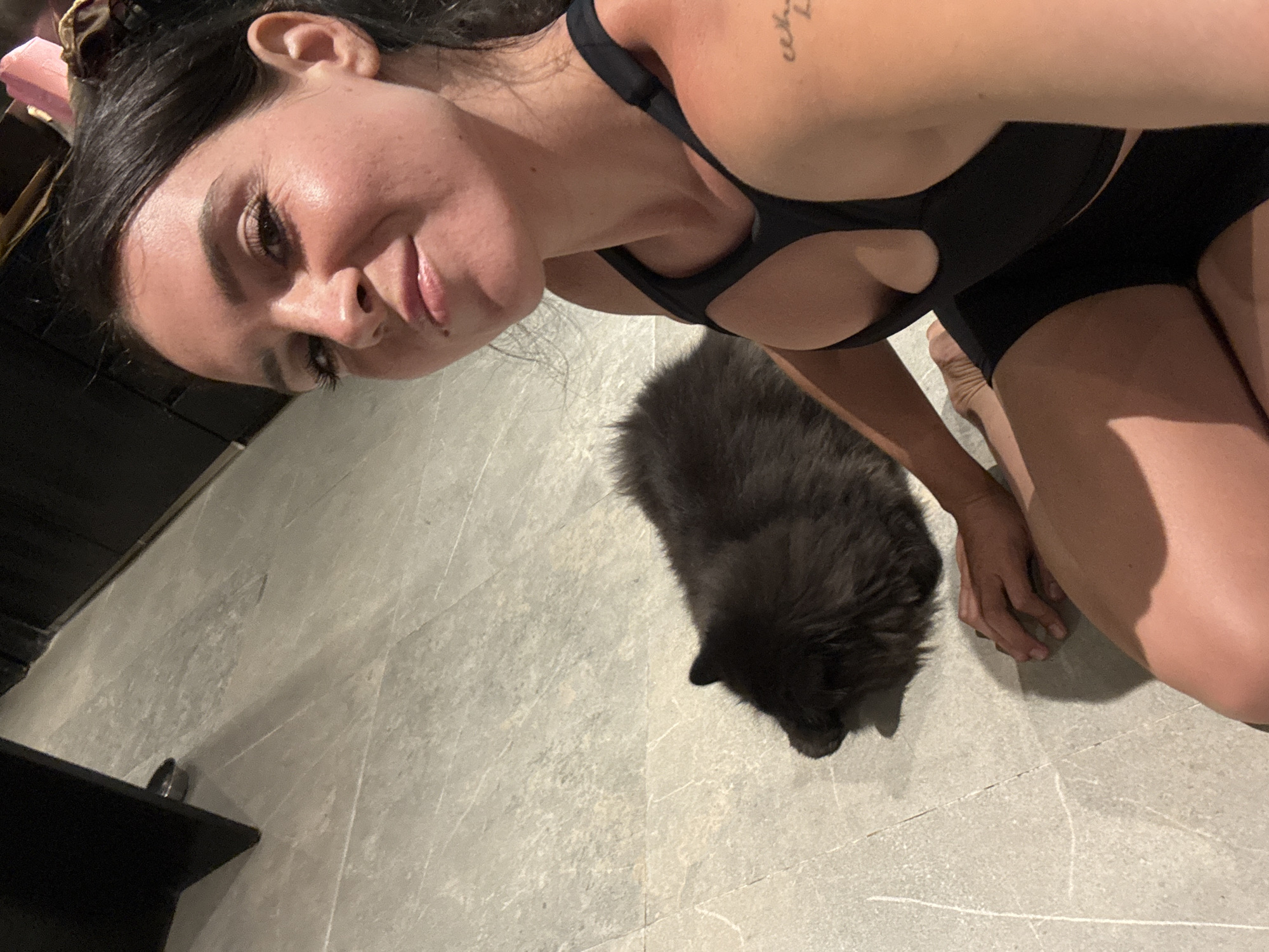 In a room with tiled flooring, a woman with dark hair is seated on the floor next to a dark-colored, fluffy cat. The woman is wearing a black sports bra and shorts, and she is taking a selfie. The cat is lying down beside her. The background is indistinct, but suggests an indoor setting.