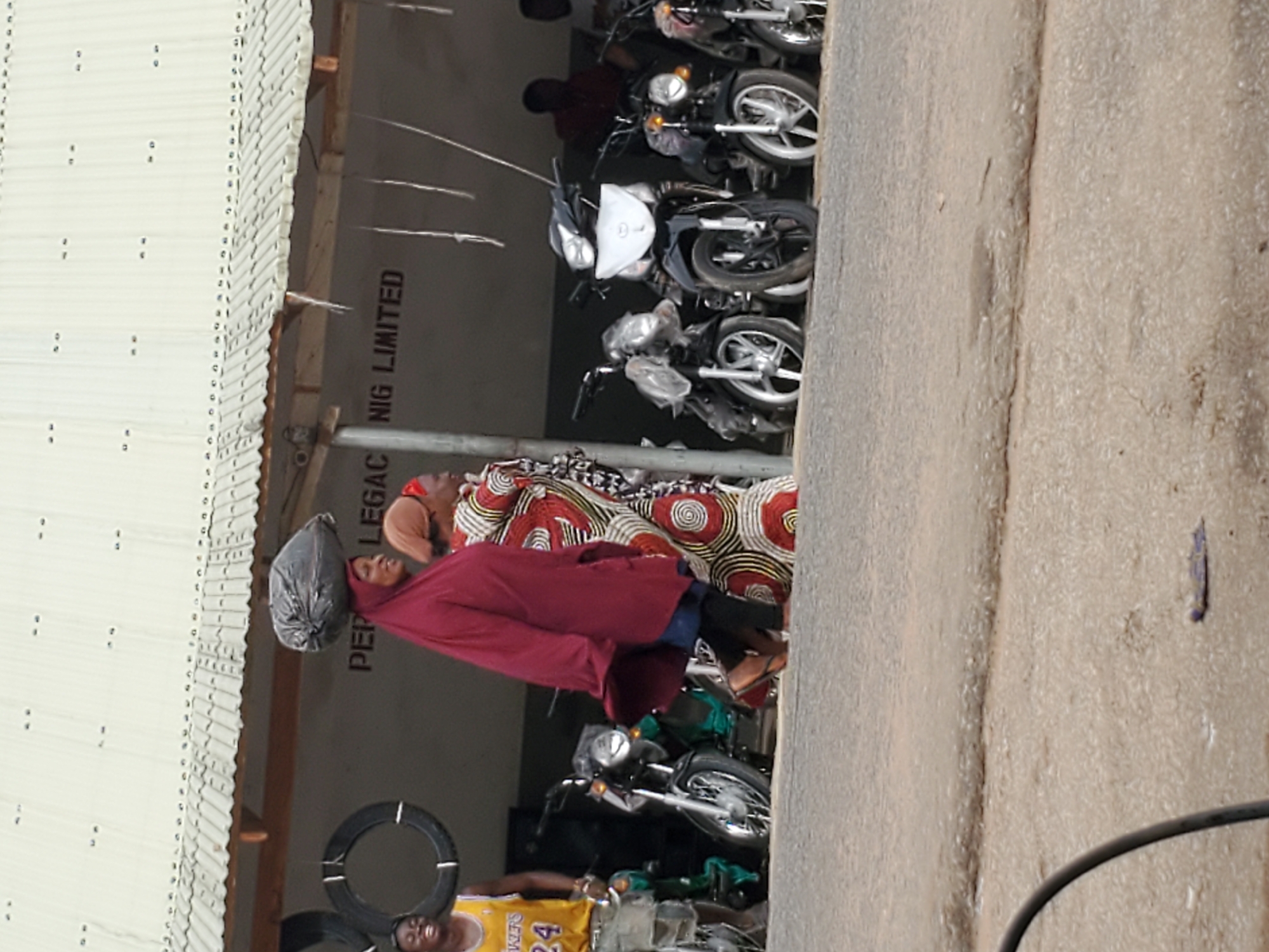 Here's a description of the image:
The image appears to be taken outdoors, possibly in a market area or roadside location in Mararaba, Nigeria. A woman with a black bag on her head is walking, wearing a red robe. Behind her, another woman is walking, wearing a colorful red-and-white patterned garment. In the background, a line of motorcycles is parked near a building. The building has a white corrugated metal roof. The text "PEP LEGAC NIG LIMITED" is visible on the wall of the building.
In the foreground, a man wearing a yellow Lakers jersey with the number 24 can be partially seen. There are tires hanging from the building and several other motorcycles in the immediate area. A segment of the road can be seen on the right-hand side of the frame. The image is slightly rotated.