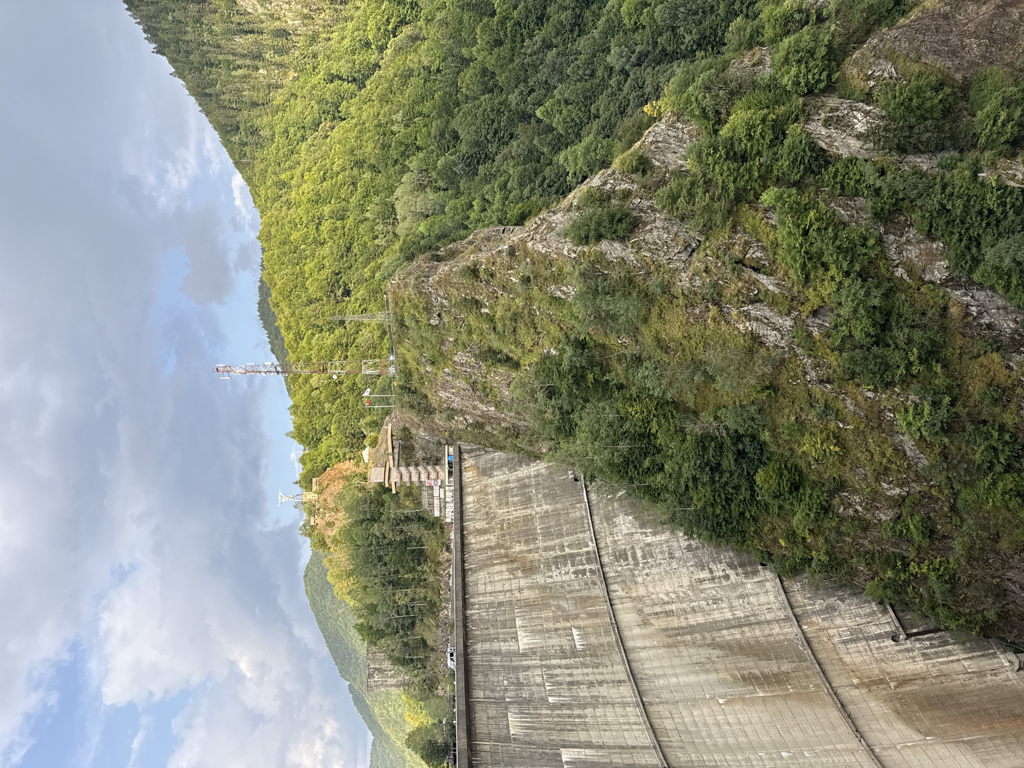 The image captures a view of the Vidraru Dam, a prominent arch dam located in Arefu, Romania. The massive concrete structure of the dam dominates the left side of the frame, its textured surface showing signs of age and weathering. A road runs along the top of the dam, with several vehicles visible, indicating ongoing traffic.
To the right of the dam, a steep, rocky cliff face covered in lush green vegetation rises dramatically. The trees clinging to the slopes are dense, creating a striking contrast with the starkness of the dam. Perched on top of the cliff is a telecommunications tower, its metal lattice structure reaching towards the sky. Further to the right, the rugged terrain continues, covered by a dense forest that stretches up the mountainside.
The sky above is partly cloudy, with patches of blue interspersed with dramatic grey clouds, suggesting dynamic weather conditions. The overall scene depicts a powerful man-made structure integrated into a wild and mountainous natural landscape.
