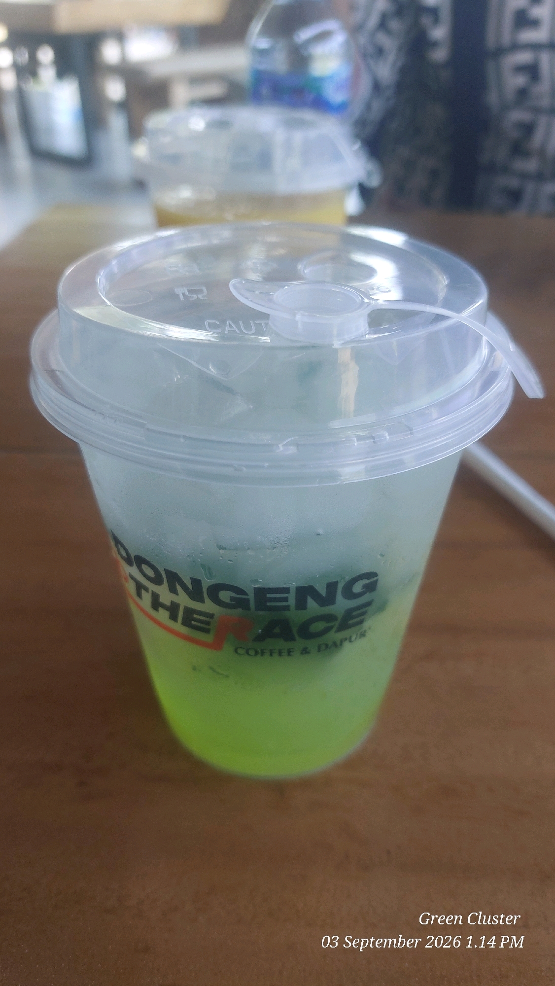 A close-up shot focuses on a tall, clear plastic cup filled with a vibrant green beverage and ice. Condensation is visible on the exterior of the cup. The cup has a clear plastic lid with a tab, and the text "CAUT" is embossed on the lid. The lower portion of the cup displays text in black and orange, with "ONG" and "ACE" being clearly legible, and the words "THE" and "R" are partially visible. Below this, the text "COFFEE & DAPUR" is written in smaller font. The cup is positioned on a wooden surface. In the background, two other cups with clear lids are visible, one containing a yellowish liquid, and beyond that, blurry figures and a bottle of water. The bottom right corner of the image contains text indicating "Green Cluster" and "03 September 2026 1.14 PM." Based on the provided metadata, the image is associated with the city of Garut, Indonesia.