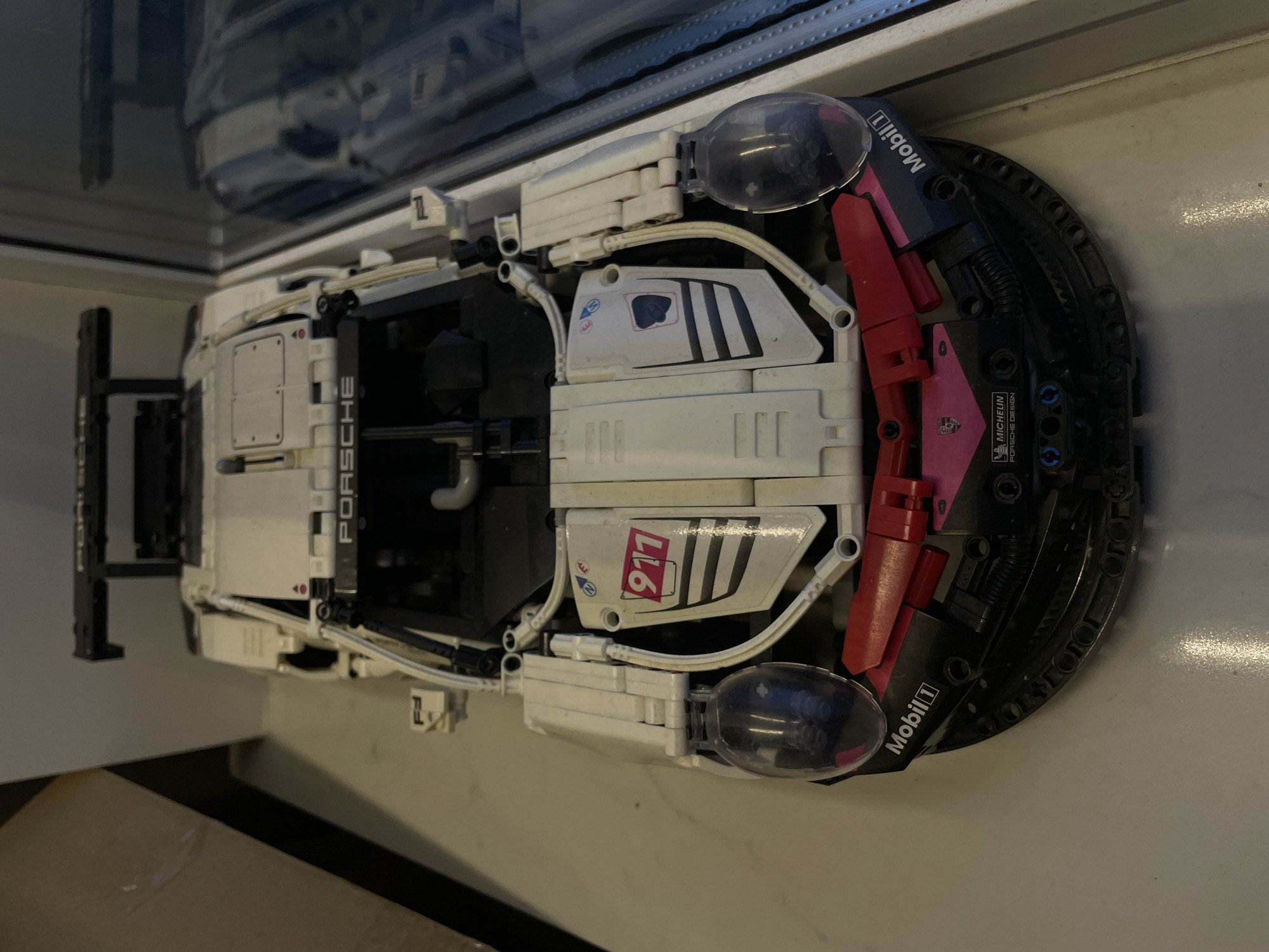 A white and black Lego Technic Porsche 911 RSR model car is displayed on a light-colored surface, possibly a table or shelf, in an indoor environment. The car is viewed from a slightly elevated, front-facing perspective. It features detailed branding, including "PORSCHE" across the windshield, a "911" graphic on the hood, and "Mobil 1" decals on the sides. The car has a large rear spoiler and visible internal mechanisms characteristic of Lego Technic sets. The background shows a blurred area that suggests a wall or partition to the left and a reflective surface, likely glass, to the right, which shows a distorted reflection of what appears to be more of the car or similar objects. The lighting is somewhat dim, creating shadows. The specific location within the city of Kommunarka, Russia, cannot be determined from the visual information provided in the image.