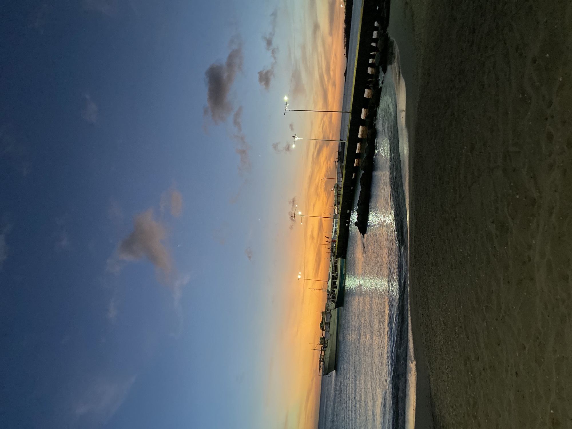 The image depicts a serene twilight or sunset scene in Juan Griego, Venezuela, overlooking a body of water that appears to be a bay or harbor. The sky dominates the upper portion of the frame, showcasing a dramatic gradient from deep indigo blue at the top to vibrant orange and yellow hues near the horizon, indicating either sunrise or sunset. Wispy, dark clouds are scattered across the sky, particularly on the bluer side. A faint, bright white dot, likely a star or planet, is visible in the upper-left blue expanse.
A long pier or seawall stretches horizontally across the middle ground, separating the colorful sky from the water. Several streetlights or pier lights are illuminated along this structure, casting bright reflections onto the calm water below. Moored boats, including what appear to be fishing vessels, are docked alongside the pier in the lower-central part of the image, silhouetted against the shimmering water. The water itself is dark but reflects the sky's colors and the piercing light from the lamps, creating a textured surface. To the right of the water and extending into the foreground, a dark, sandy or muddy shoreline completes the coastal setting. The overall scene evokes a peaceful and tranquil atmosphere at the transition from day to night. No specific human activity or text is visible.