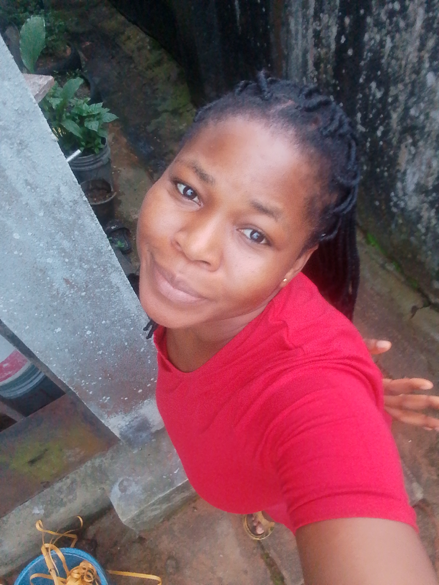 A high-angle, medium shot shows a woman in a red t-shirt, standing outdoors. She has dark skin, braided hair, and is looking up at the camera. Her right hand is extended towards the camera. In the background, there is a rough, grey, concrete wall and some greenery. To the left of the woman, there is a grey concrete railing, a blue bucket with yellow straps, and a section of what appears to be a painted cylinder. The ground is a mix of concrete and dirt. The scene appears to be located in an urban outdoor environment, possibly in Yenegoa, Nigeria, given the provided context.
