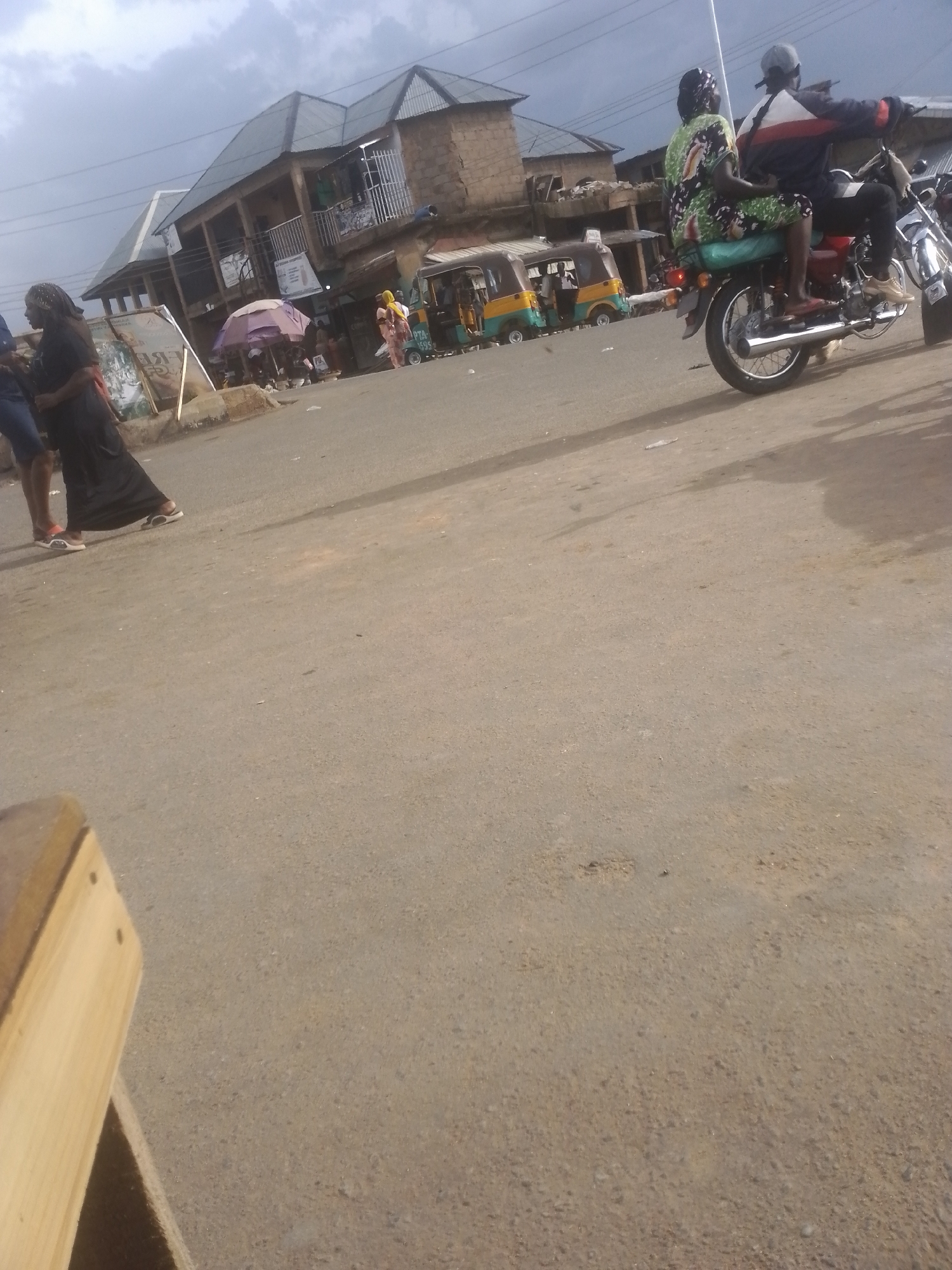The image depicts a street scene, likely in a marketplace. In the foreground, a portion of a wooden bench is visible on the left. The road surface is paved and appears somewhat worn. In the middle ground, several people are present. On the left, a woman in a long black dress and white sneakers is walking. Further into the scene, there are shops and stalls lining the street. A purple umbrella is set up, and several individuals are gathered around. A yellow and green three-wheeled vehicle, resembling a tuk-tuk, is parked on the right side of the street, with another similar vehicle further behind it.
To the right of the scene, a motorcycle carrying two people is moving away from the viewer. The rider is wearing a grey cap and a jacket with red and white stripes. The passenger, a woman, is wearing a colorful patterned dress and a headscarf.
The background shows a row of buildings, some with multiple stories and corrugated metal roofs. The sky is overcast with clouds, suggesting it might be daytime, perhaps late afternoon, with diffused light. The overall atmosphere appears to be that of a busy, everyday scene in a commercial area. No discernible text is clearly visible in the image.
