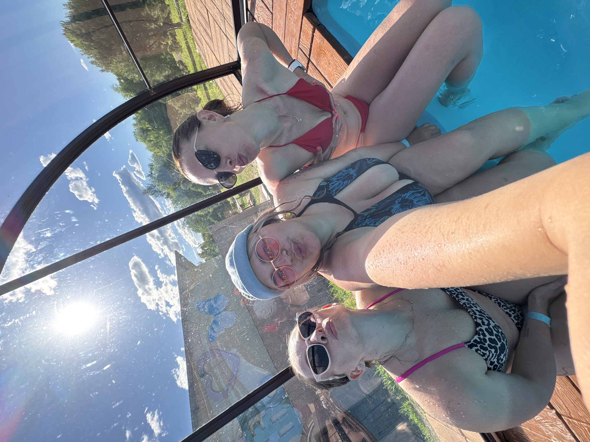 Three individuals are taking a selfie in a small, enclosed pool on a sunny day. The pool is surrounded by a wooden deck and is covered by a transparent, arched roof. The individuals are wearing swimsuits and sunglasses, and their bodies are wet, indicating they are in or have recently been in the water. The person in the center is wearing a blue hat and pink-rimmed sunglasses, and is making a kissing face. The person on the left is wearing leopard-print swimwear and pink-rimmed sunglasses, also making a kissing face. The person on the right is wearing a red bikini and black sunglasses, and is touching their chest. The background shows greenery and trees, suggesting an outdoor setting. The image is bright, with the sun shining through the roof, creating glare and reflections on the transparent surface. The scene appears to be a leisure activity at a resort or private property, possibly in a natural or semi-rural environment, though the specific location in Birobidzhan, Russia cannot be determined from the image alone.