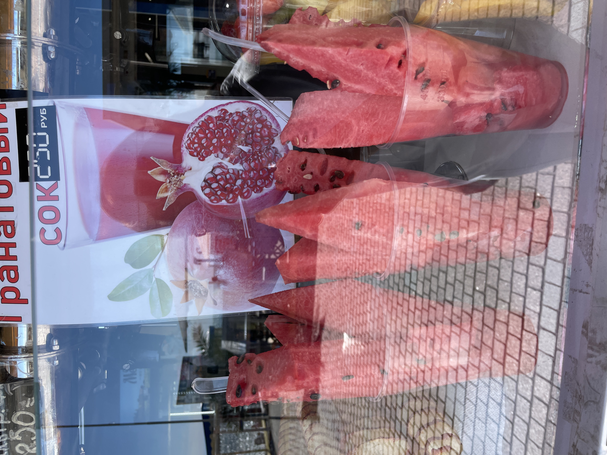 This close-up image shows a street vendor's or shop's glass display window in Sochi, Russia, filled with fresh fruit. The primary subjects are several clear plastic cups, each holding bright red, cut watermelon pieces. Some pieces have small plastic forks or spoons inserted, indicating they are ready for sale and consumption. The watermelon pieces show their characteristic dark seeds.
Behind and partly reflected in the glass, there is a poster advertising juice. Visible Cyrillic text on the poster reads "ГРАНАТОВЫЙ СОК" (Pomegranate Juice) and "250 РУБ" (250 RUB), indicating its price. The poster also features an illustration of a sliced pomegranate. Faint volume markings, "400ml" and "330ml," are visible on one of the plastic cups.
The scene is brightly lit by daylight, suggesting it is daytime. Strong reflections on the glass surface obscure parts of the display and reveal external elements, including a patterned pavement or cobblestone street and indistinct architectural features in the background. The display of fresh fruit, particularly watermelon, in such a setting suggests a warm climate or season. No people are visible, but the display is clearly prepared for customers.