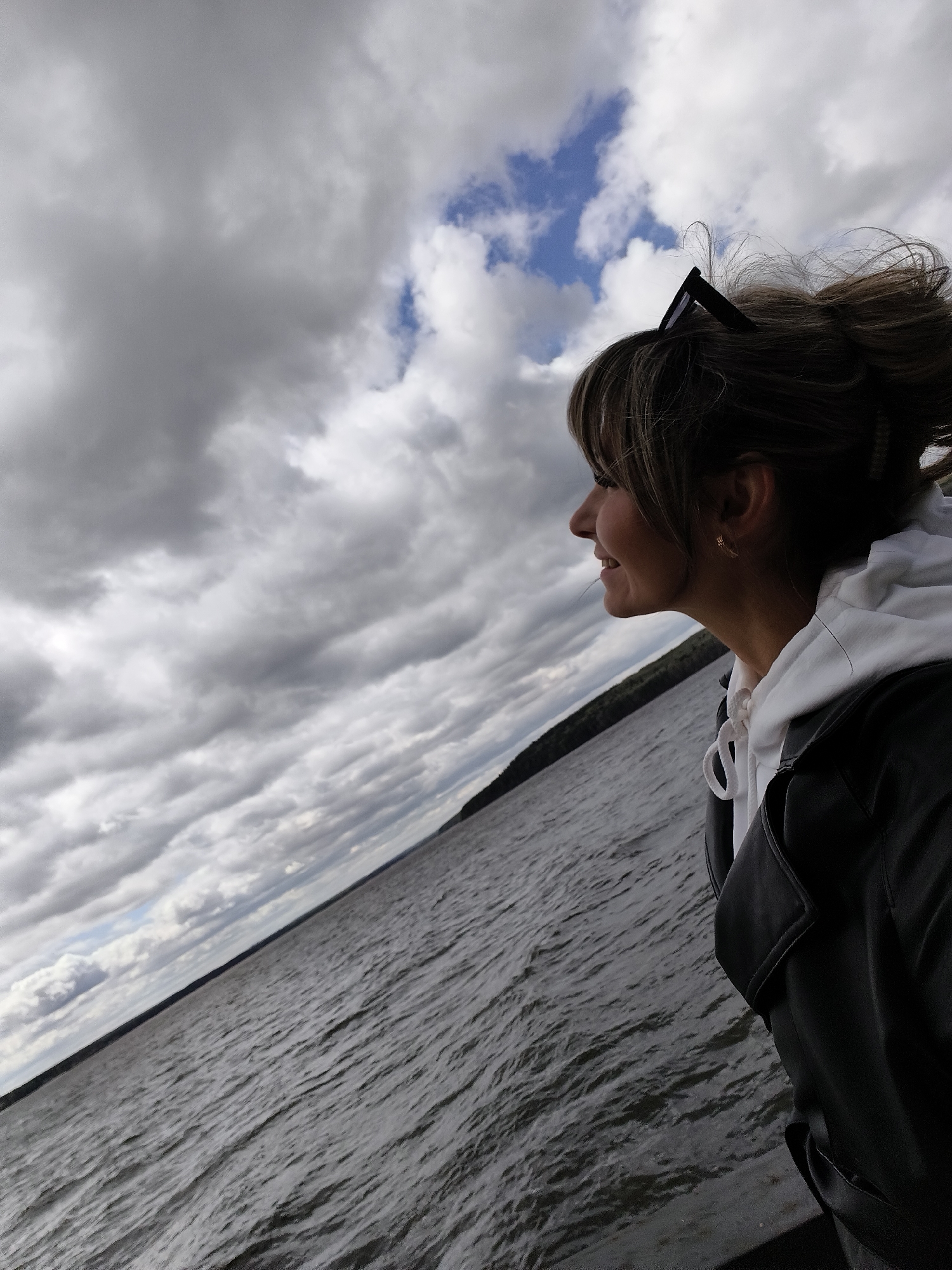 A medium, eye-level shot shows a woman looking out over a body of water under a cloudy sky. The woman has light skin, dark hair pulled back with some bangs in front, and she is wearing sunglasses on her head, a white hoodie, and a black jacket. She is in profile, facing left, and has a slight smile. The water is choppy, with visible ripples. In the distance, there is a strip of green trees along the horizon. The sky is dominated by gray clouds, with patches of blue visible. Given the information provided, the location is Chaikovsky, Russia.