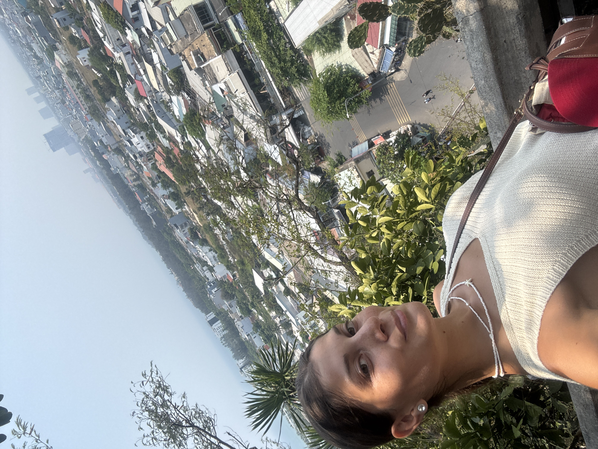 A woman is photographed against a backdrop of an urban area in Đà Nẵng, Vietnam. The city is visible with houses and streets, and the sea in the distance. The woman is wearing a white top and a necklace. A red and brown purse is visible beside her. She is standing near trees and greenery.