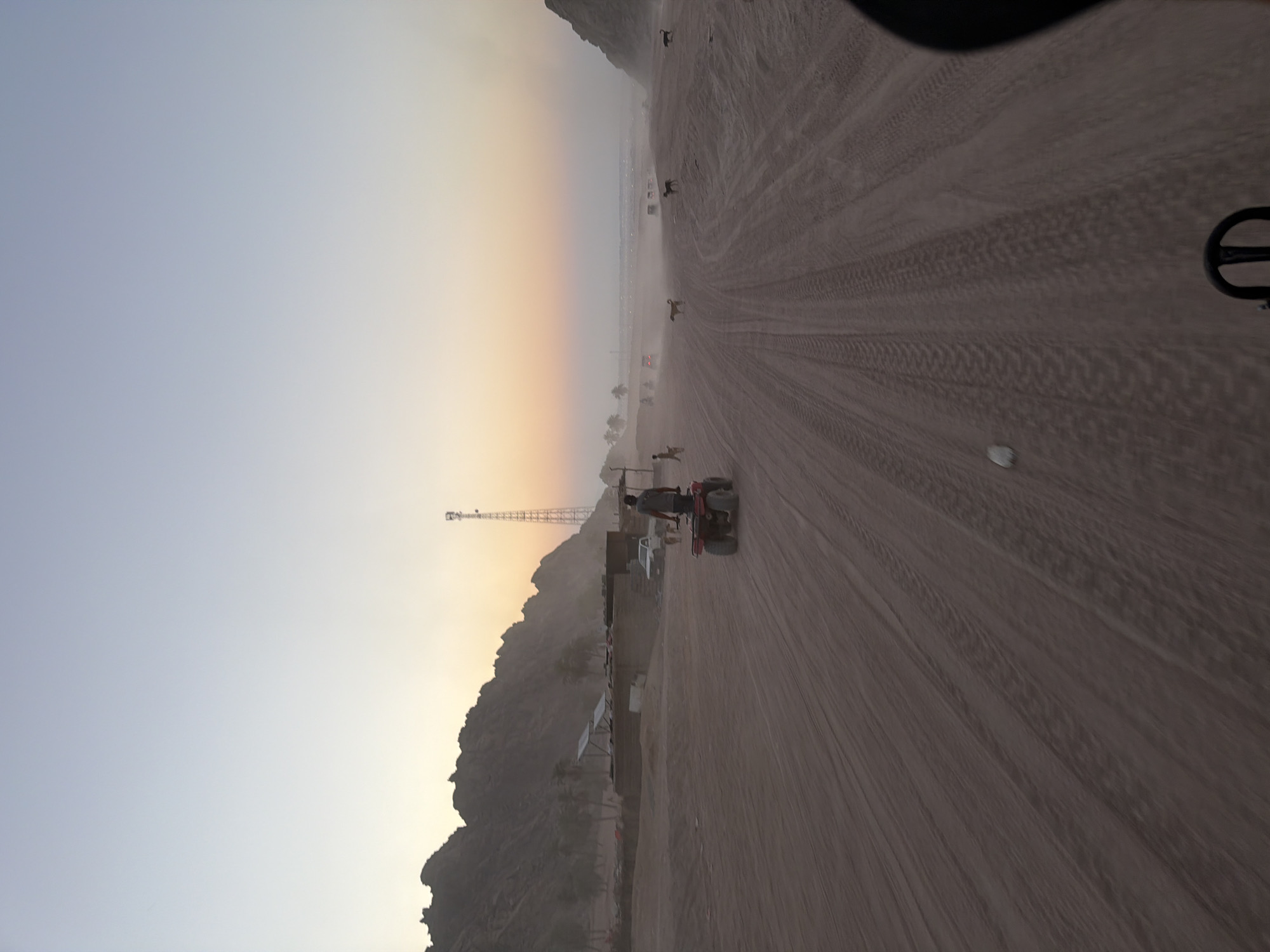 This image depicts a dusty desert landscape near Sharm El Sheikh, Egypt, likely during sunset. In the foreground, a person is riding an all-terrain vehicle (ATV) on a sandy track marked with tire imprints. The ATV is red and appears to be moving away from the viewer. In the middle ground, there are some low-lying structures that resemble dwellings or temporary settlements, with solar panels visible on one roof. A white pickup truck is parked near these structures. Further in the background, several dogs are scattered across the sandy terrain. The horizon is hazy, with a tall telecommunications tower visible against the gradient sky, transitioning from a pale blue at the top to warmer tones of orange and yellow near the horizon, indicating the setting sun. The mountainous terrain forms a dark silhouette on the left side of the image. The overall atmosphere is arid and sun-baked.