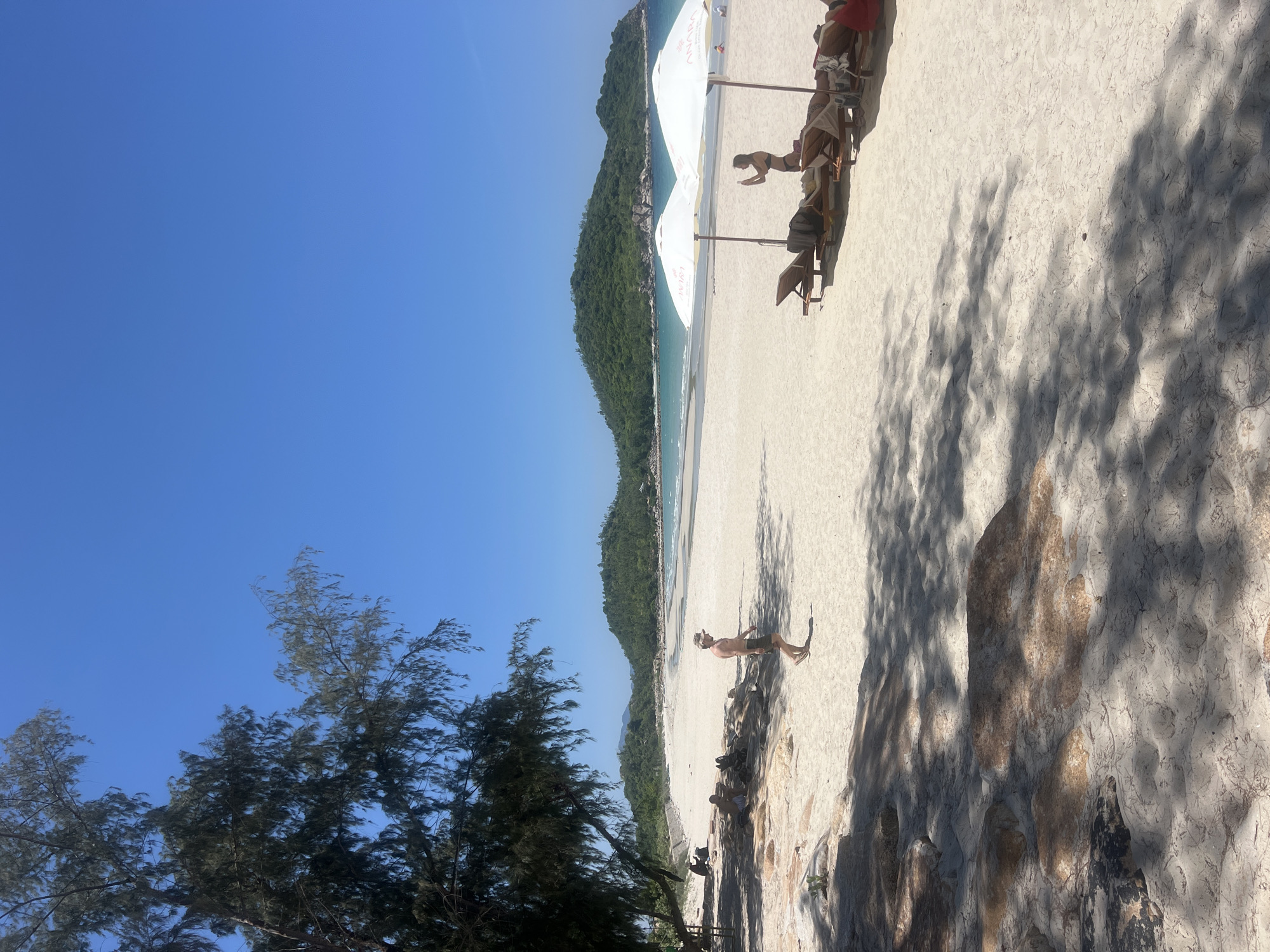 The image depicts a sunny day at a beach, likely in Cong Hai, Vietnam. The foreground is dominated by light-colored sand with scattered rocks and tree shadows. To the left, a man in swim trunks and a hat walks across the sand. Further back, near the water's edge, a few people are seated on rocks under trees. In the mid-ground, a row of lounge chairs is set up on the sand, partially shaded by two white umbrellas. A woman in a bikini stands by the lounge chairs, holding a phone, possibly taking a picture. The background features a calm, blue sea meeting a white sandy beach, with lush green hills in the distance. The sky is clear and blue.