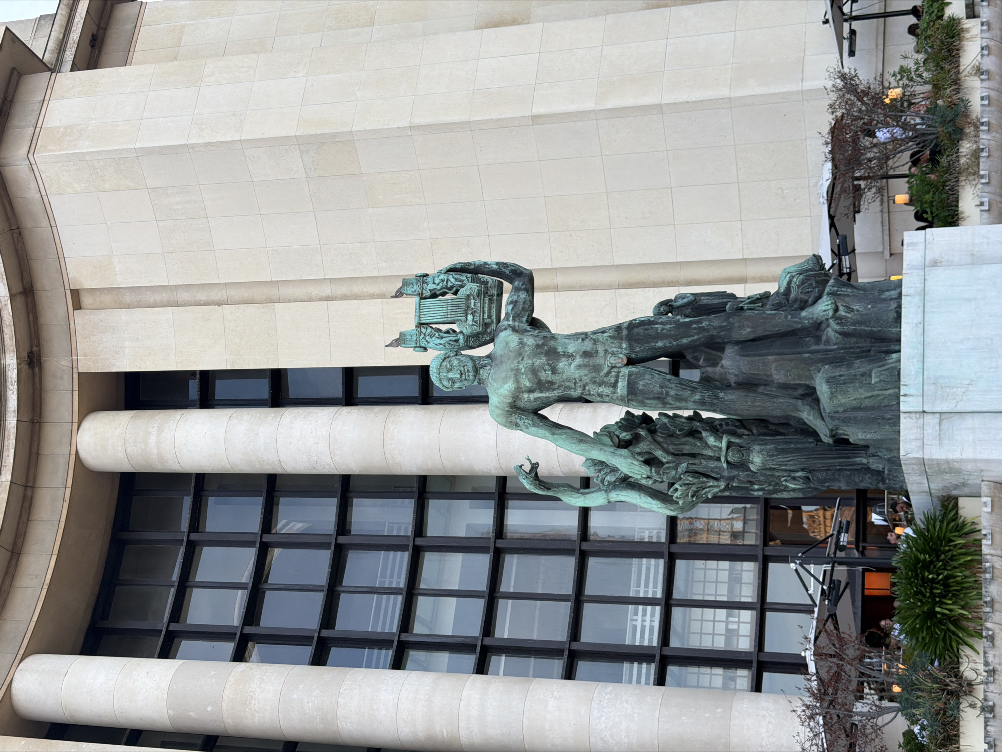 Here's a detailed description of the image:
The image presents a statue in front of a building in Paris, France. The statue is of a male figure holding a lyre or harp-like instrument above his head. It appears to be made of bronze or another dark-colored metal, and there are visible signs of weathering or oxidation, giving it a green or bluish-green hue. The statue is mounted on a sculpted base that depicts figures or forms, suggesting a dynamic and expressive scene.
The building behind the statue has a modern architectural style. It features a symmetrical facade composed of horizontal bands of light-colored stone or concrete. The building also has sections with large, dark-framed windows that form a grid pattern. There are architectural details like curved walls or columns, which add visual interest to the building's design.
Additional elements in the scene include landscaping with various plants and what appear to be decorative lighting fixtures on the right side of the image. The scene appears to be well-maintained and located in a public or prominent area of Paris.