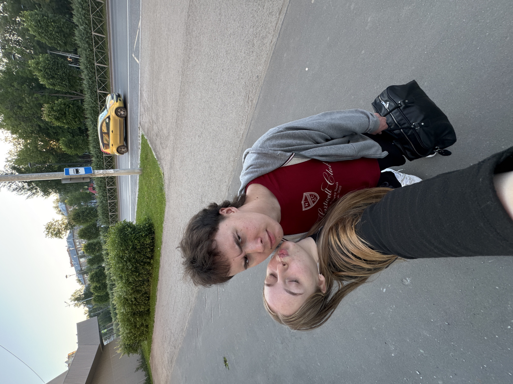 A selfie is taken from a low angle of a young couple standing on a paved area. The woman in the foreground is leaning into the frame, puckering her lips, with her eyes closed. She has long, light brown hair with blonde highlights. She is wearing a black long-sleeved top and dark pants. The man is behind her, looking directly at the camera with a stern expression. He has dark, layered hair, a mullet, and is wearing a red t-shirt with a crest on it and a grey hooded jacket. He is carrying a black duffel bag. In the background, a yellow car drives on a road. There is a bus stop sign on a pole. The scene appears to be outdoors in a city, possibly near a park or a street lined with trees and buildings. The sky is clear and the lighting suggests it is daytime. The context of the image is not provided, but the pose and expression of the couple suggest a lighthearted or affectionate moment. The environment suggests a public urban space. The presence of a bus stop and road indicates a transportation hub or a street in a developed area.
