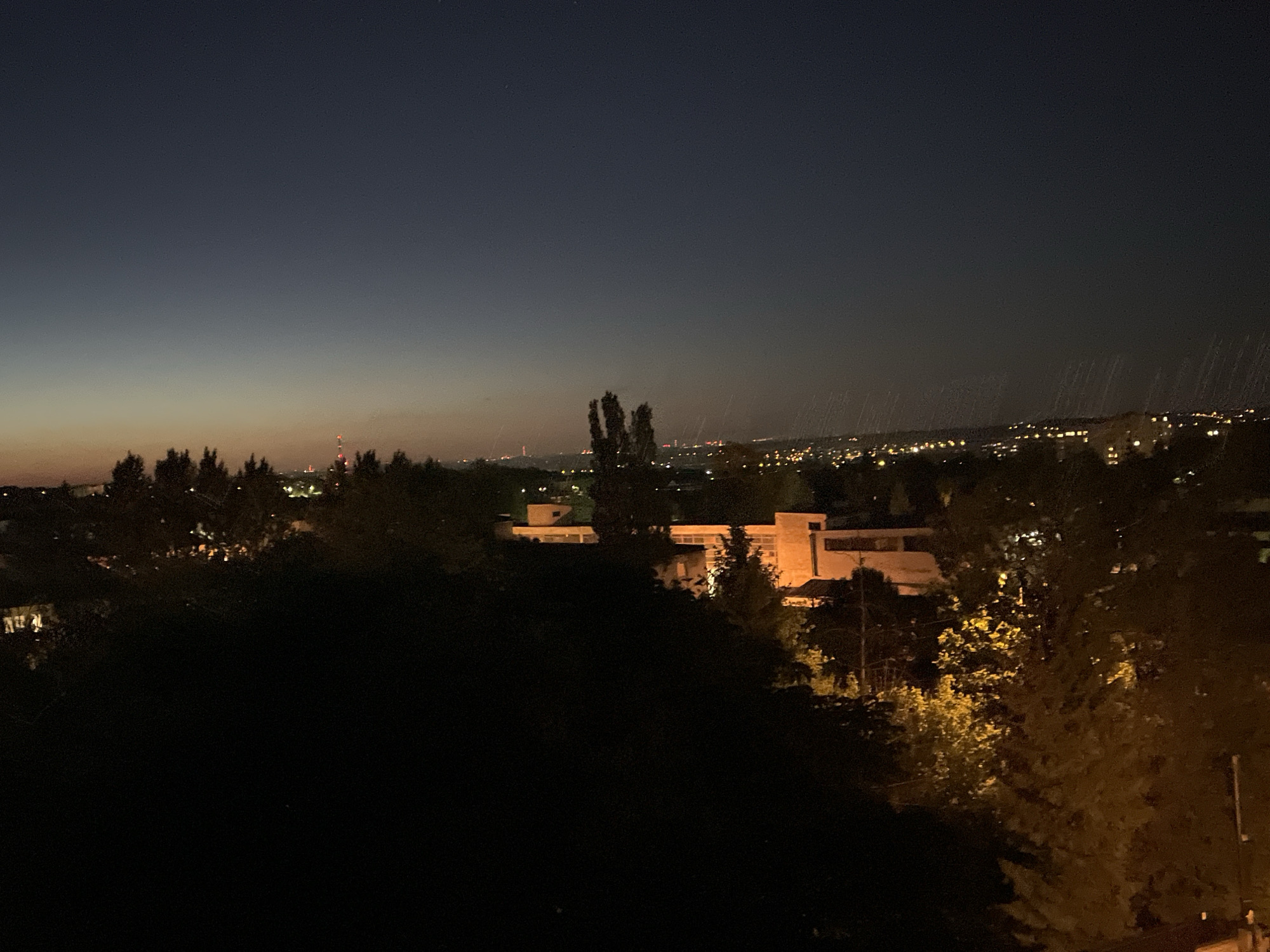 The image shows a nighttime urban view, with a building complex illuminated in the center. The foreground is dominated by dark foliage, obscuring the view partially. In the distance, city lights stretch across the horizon, indicating a dense urban area. The sky transitions from a dark orange near the horizon to a deep blue/black. There are some visible rain showers in the distance above the city lights. A structure in the middle distance is lighted up. A communications tower is visible on the left.