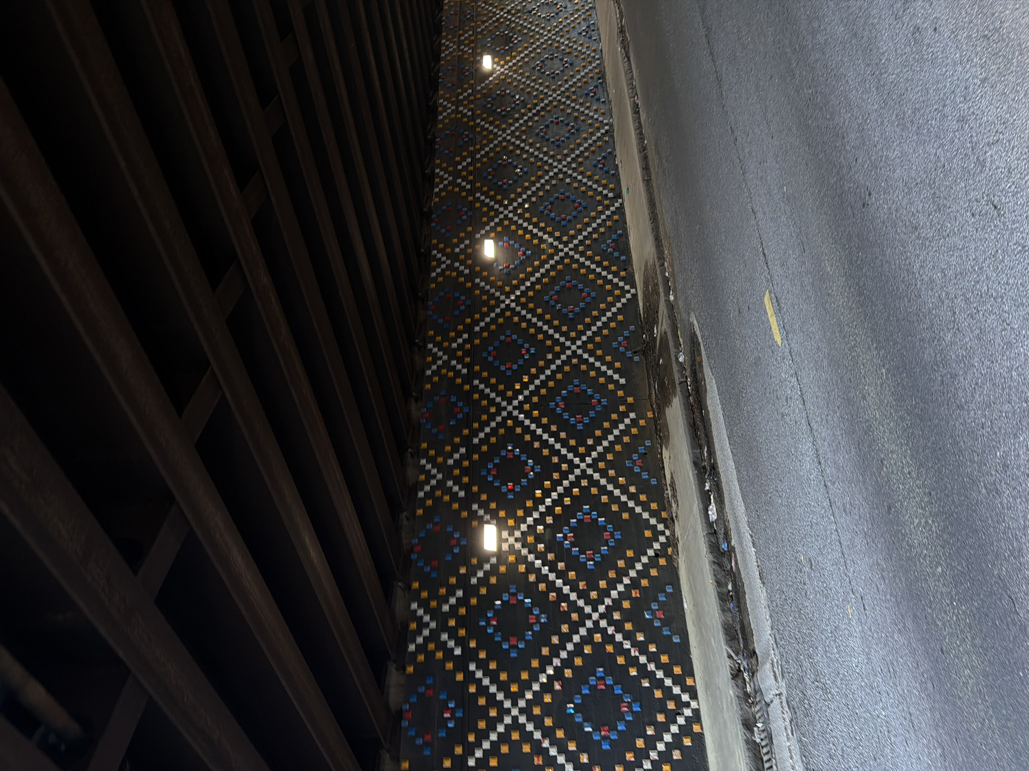 The image shows a section of the Jackson Street Bridge in Atlanta, Georgia. A patterned barrier separates a parking garage from the bridge's roadway. The barrier features a dark background with a repeating diamond pattern composed of small, colorful squares in blue, white, yellow, and red. Embedded lights illuminate the patterned barrier. To the left, the dark structure of the parking garage is visible, with horizontal beams creating a grid-like appearance. To the right, the gray asphalt of the bridge roadway is seen. A yellow stripe is partially visible.