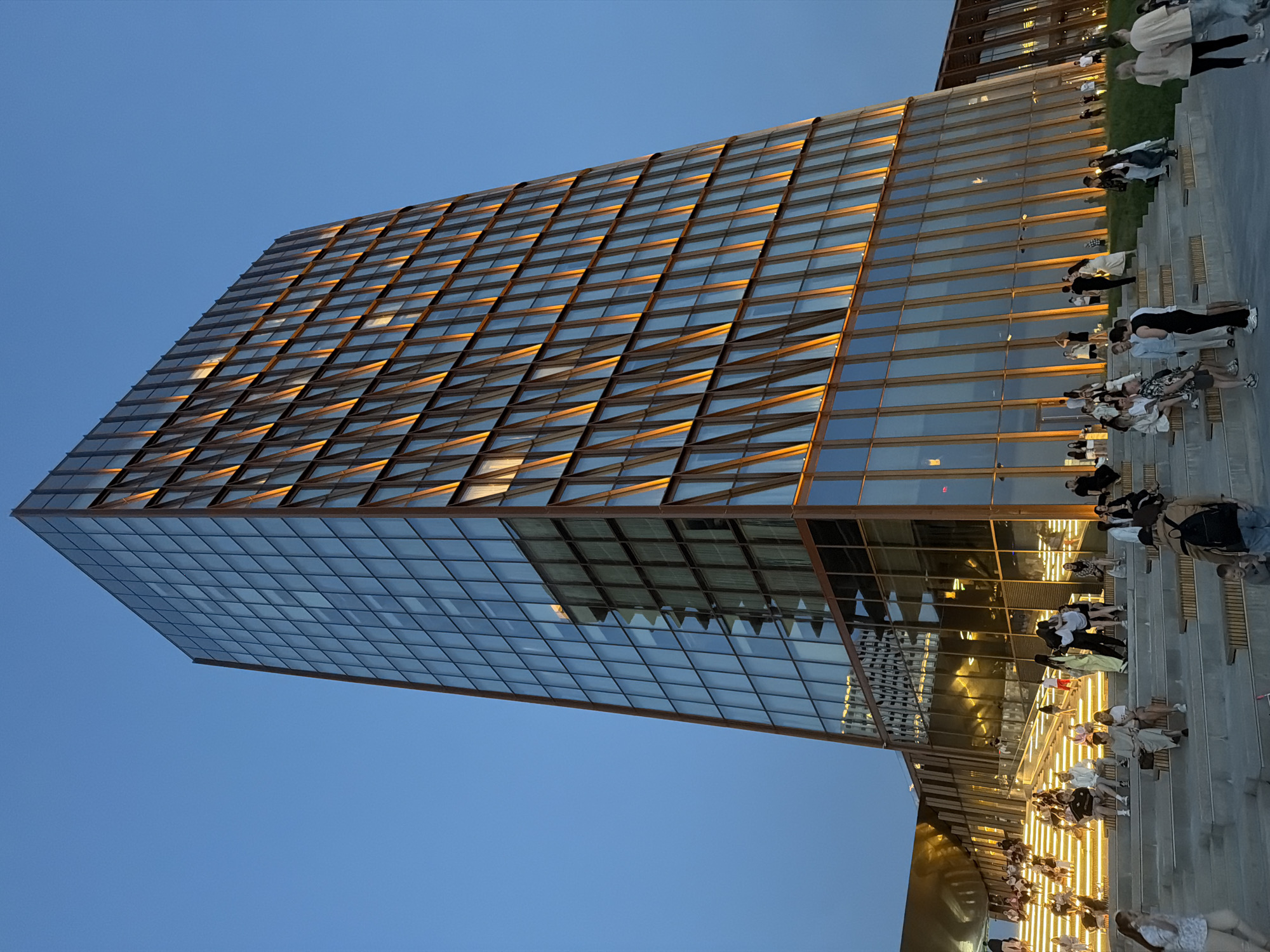 The image shows a modern building with a grid-like facade in Kazan, Russia. The building's exterior consists of numerous rectangular glass panels framed by metal. Some of the panels reflect a warm, golden light. The structure rises against a clear blue sky. In the foreground, there are people walking and sitting on steps or benches. The people are dressed in casual clothing. The overall scene suggests a public space adjacent to the building.