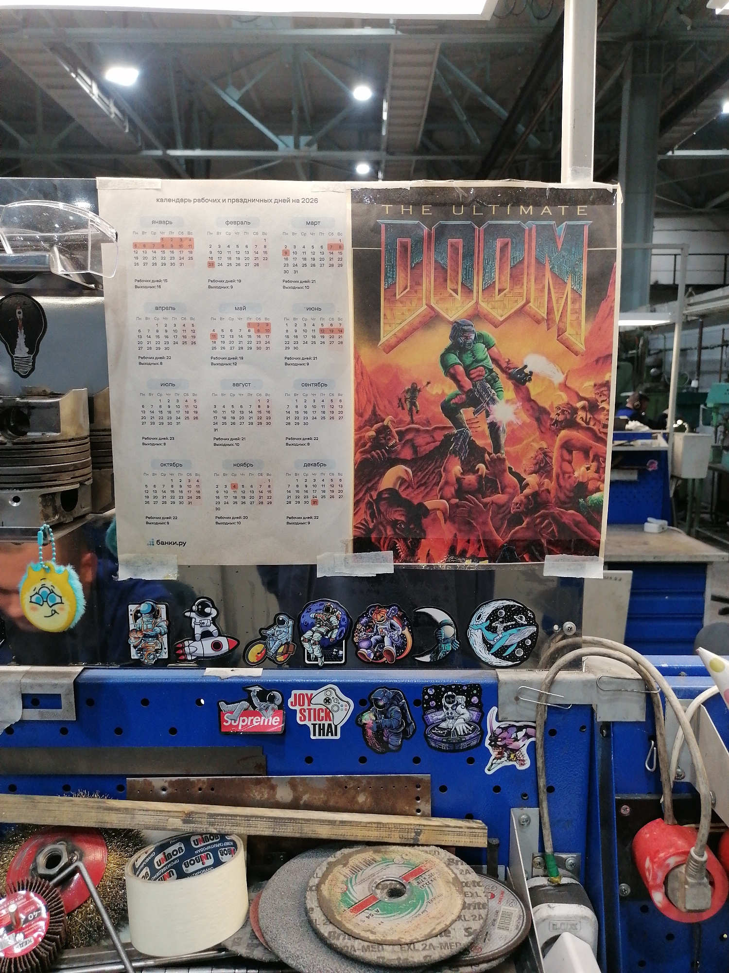 A cluttered workbench in an industrial setting features a 2026 calendar (in Russian) and a "The Ultimate Doom" poster taped to the front of a blue metal shelving unit. The shelves are adorned with a variety of stickers, including space-themed images and brand logos like "Supreme" and "Joy Stick That." Various tools and materials, such as grinding discs, wire brushes, and tape, are scattered on the shelf below. A yellow plush keychain with a multi-eyed design is hanging on the left. The scene appears to be a workspace where tools and personal items are mixed.