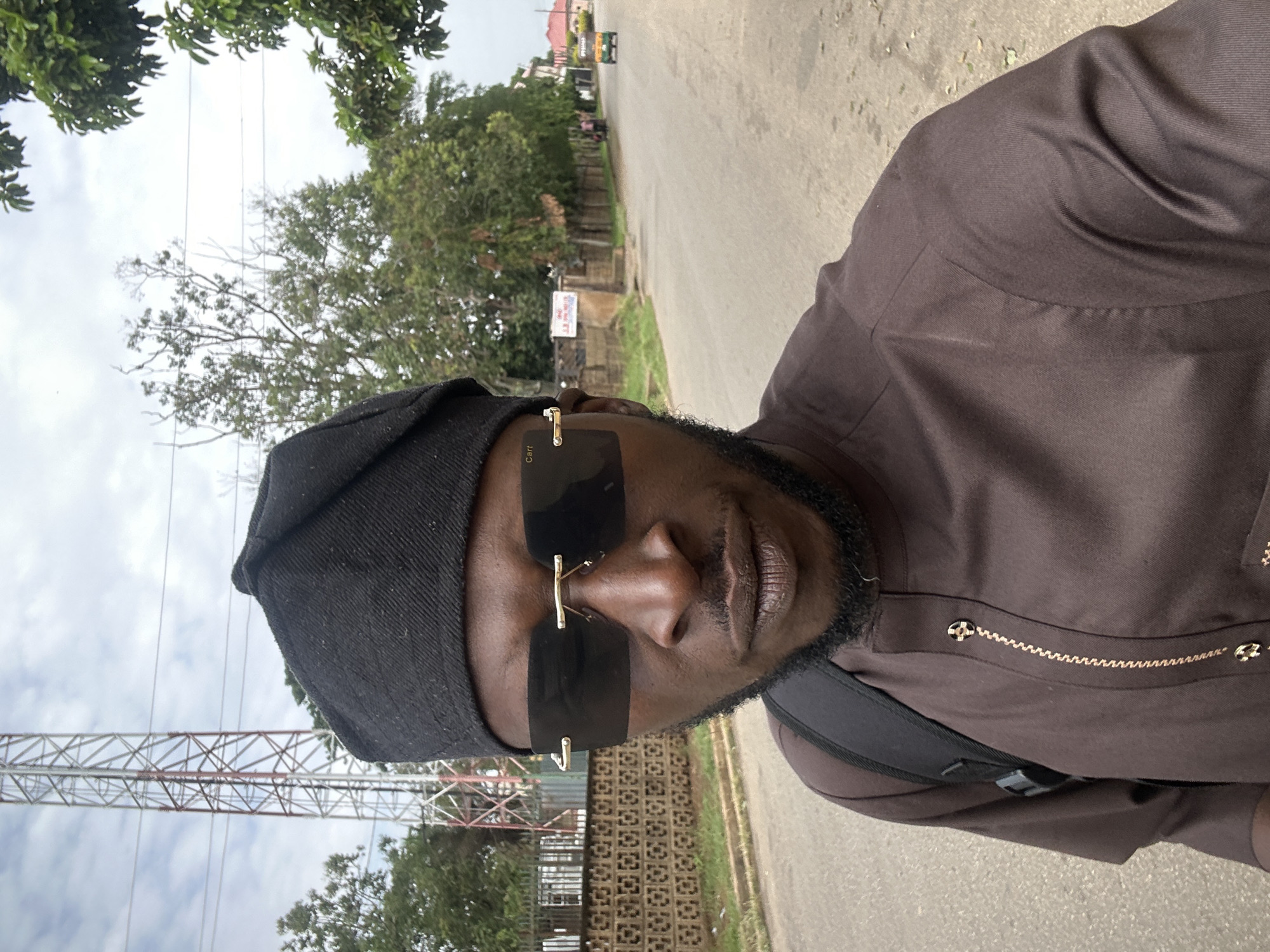 A person is standing on a street in Jos, Nigeria, as suggested by the architecture and environment. The individual is wearing a dark brown tunic and a black cap, sunglasses, and appears to be wearing a backpack. The street runs away from the viewer and has trees and low buildings along its sides. An electricity transmission tower is visible to the left.