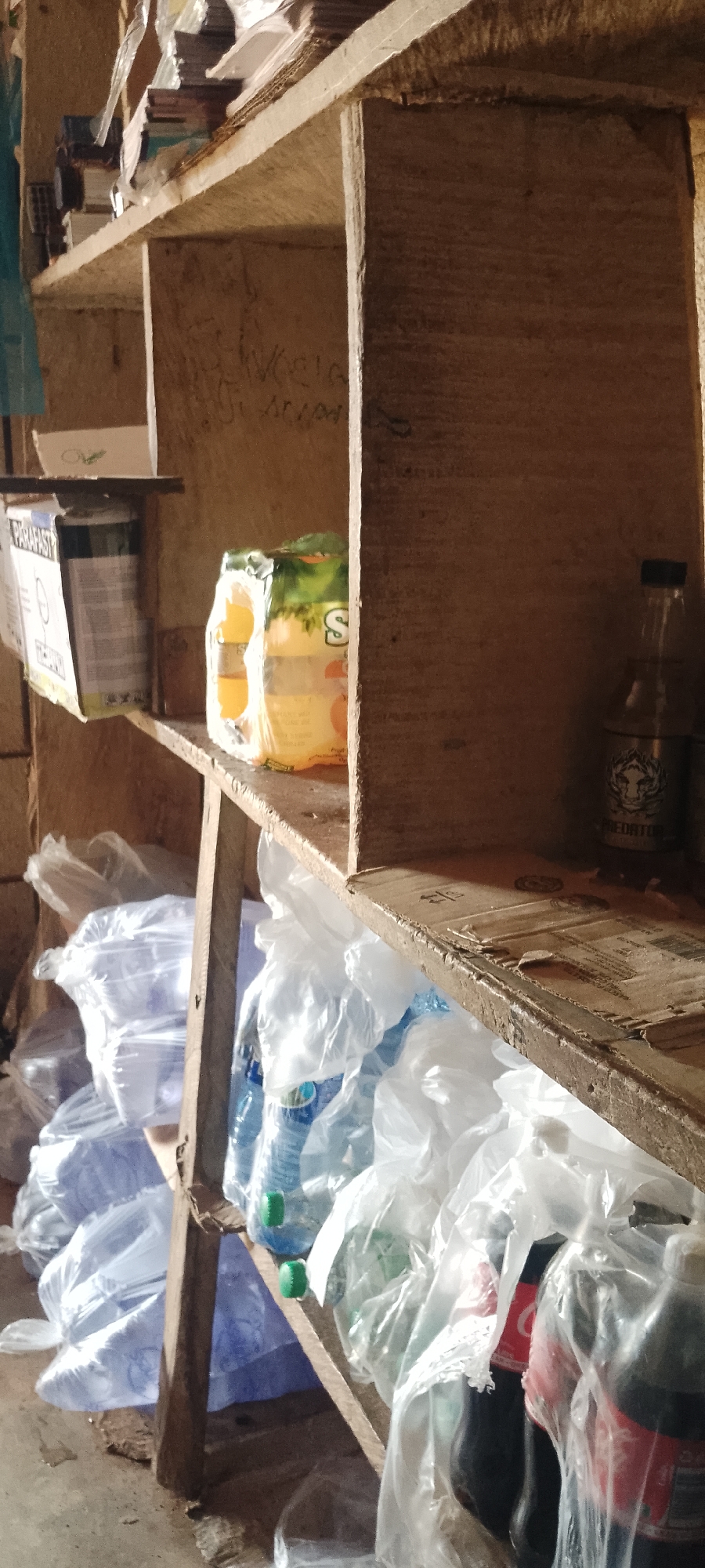 The image depicts a rustic wooden shelving unit displaying various products. On the top shelf, there are stacks of what appear to be books or paper goods, along with a boxed item labeled "PARAPASTE". To the right, a pack of drinks, possibly juice, is visible. The middle shelf holds a bottle with a dark liquid and a label featuring a tiger's head, identifiable as "PREDATOR". The bottom shelf is laden with numerous plastic bottles, many of which are covered in clear plastic bags, suggesting they are either being stored or transported. Some of these bottles appear to be water bottles with green caps, while others are dark-colored, resembling soda. The overall environment appears to be a storage area or a small shop, with the wooden shelves showing signs of wear and some graffiti on the back panel. The lighting suggests an indoor setting, possibly with natural light entering from the side. This scene is located in the city of Durumi, Nigeria.