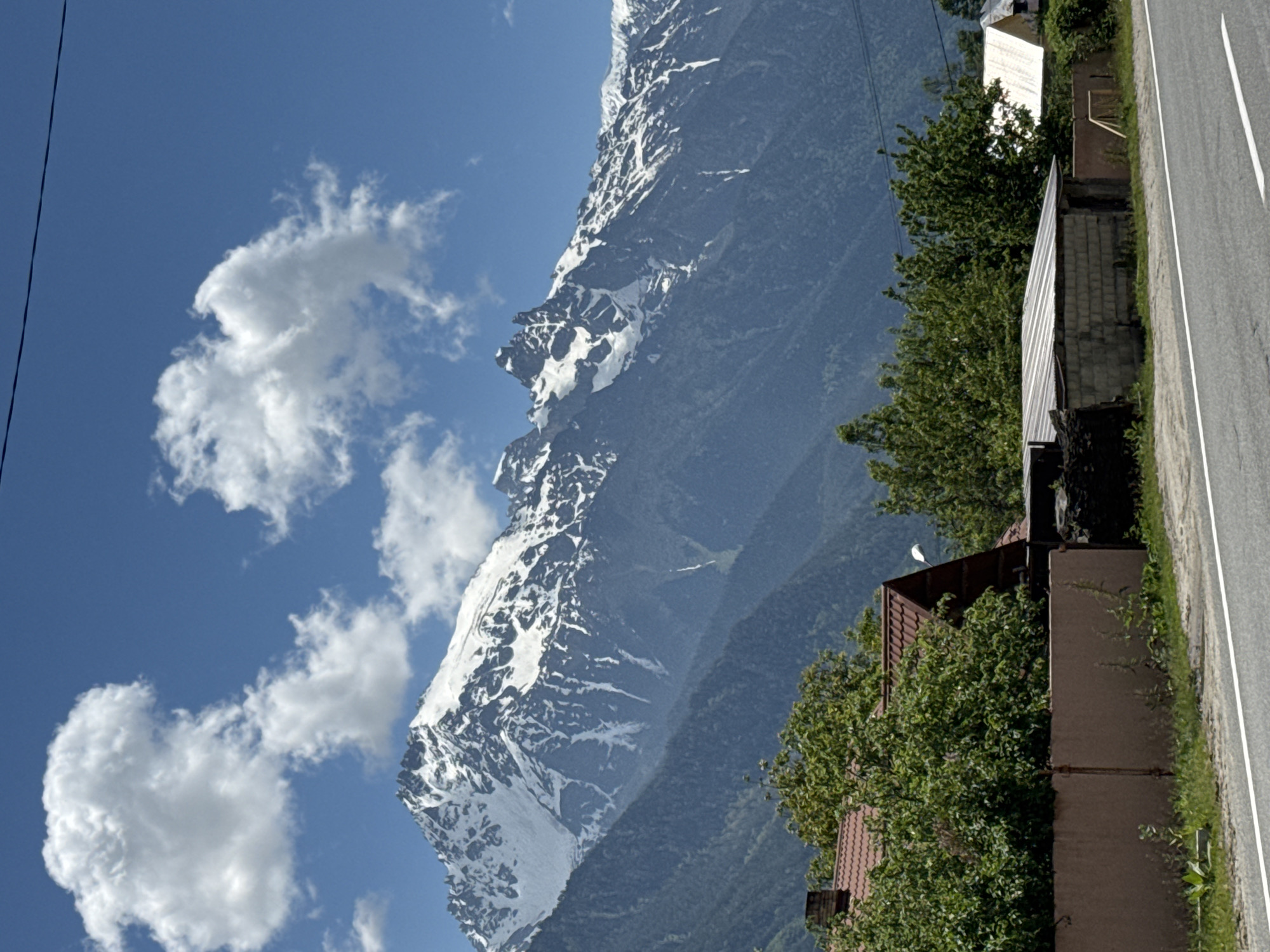 The image depicts a scenic mountain landscape with a small village in the foreground, likely located in Teberda, Russia. Dominating the background is a majestic mountain range, its peaks capped with snow, under a clear blue sky dotted with fluffy white clouds. The slopes of the mountains are covered in dense green forests. In the mid-ground, several houses with red and white roofs are visible, nestled amongst lush green trees. A brown fence runs along the edge of a road in the foreground, with a paved surface and a white line marking the lane. The scene suggests a peaceful, rural environment with a striking natural backdrop.