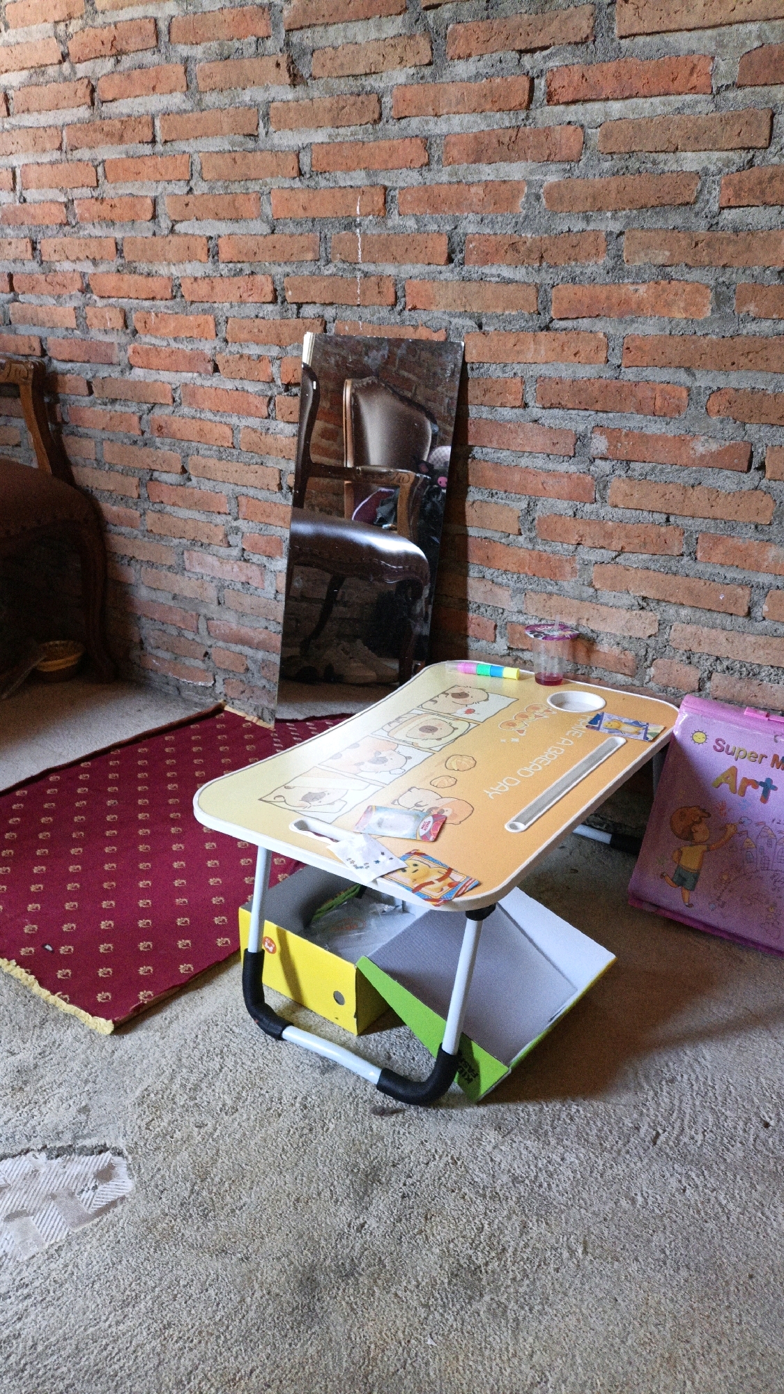 A portable lap desk with a yellow, cartoon-themed surface sits on the floor in front of a brick wall. The desk has foldable legs and is propped open with two colorful boxes underneath. A small mirror leans against the wall behind the desk, reflecting a portion of the room. To the left of the desk is a red rug with a repeating pattern. To the right, a purple book with the words "Super Master Art" is visible. The floor is a rough, concrete-like material. The overall impression is one of a casual, indoor setting, possibly a child's play area or a study space. No people are present in the image.