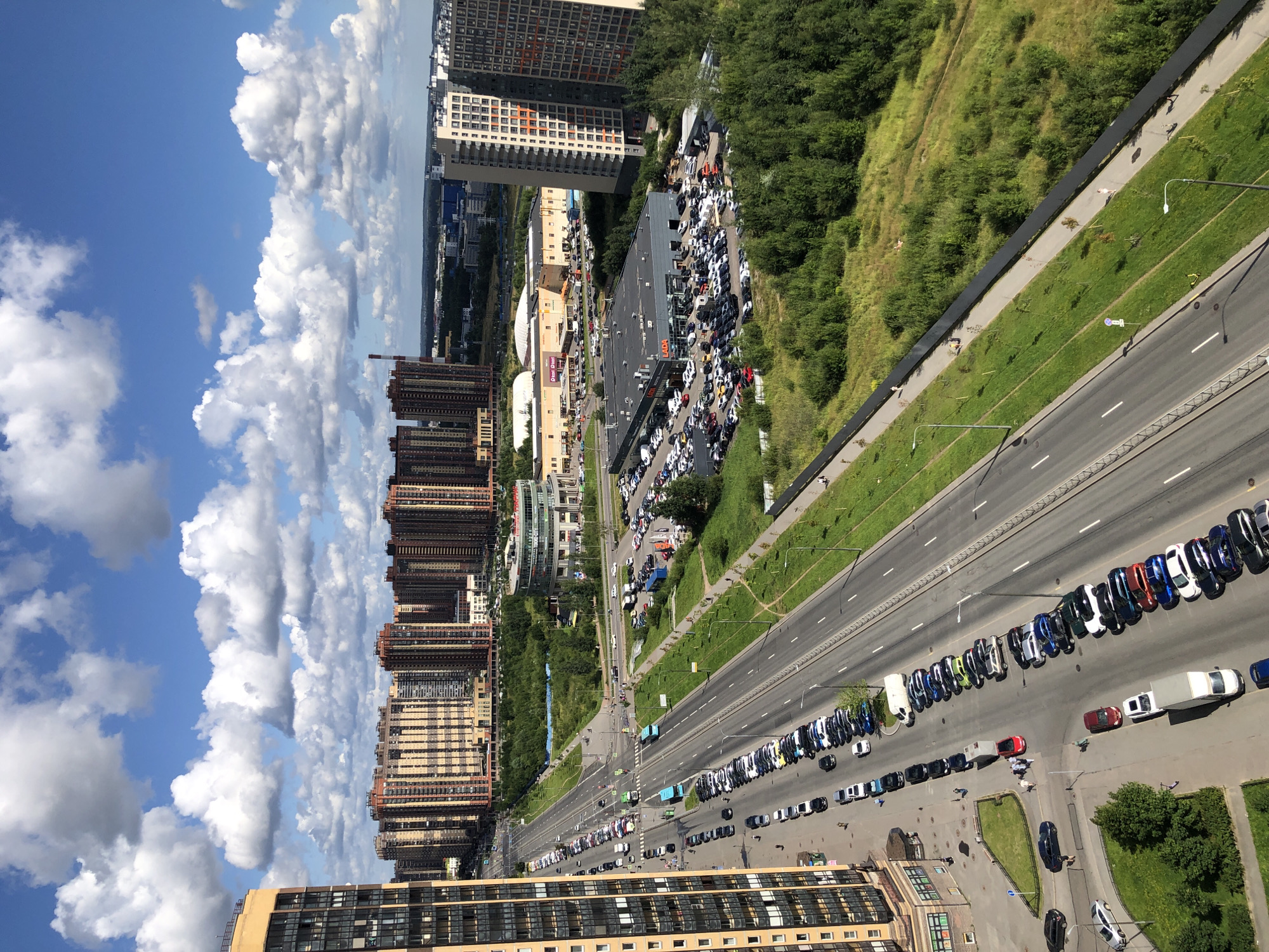 An eye-level aerial view captures a modern urban landscape under a bright blue sky dotted with fluffy white clouds. The scene is dominated by a multi-lane road running diagonally from the bottom left to the upper right of the frame. The road is lined with rows of parked cars on both sides, with a consistent stream of vehicles in motion on the lanes.
To the left of the road, a tall, modern apartment building with a beige facade and dark windows rises prominently. Across the road, several large, multi-story residential buildings with a repeating pattern of beige and reddish-brown facades create a dense urban backdrop. Further in the distance, a low-lying commercial building complex with a large circular section and a sprawling parking lot is visible. Adjacent to this, a car dealership, identified by a large "LADA" sign, is packed with vehicles.
On the right side of the road, a steep, vegetated embankment covered in lush green trees and grass slopes upwards, separated by a dark fence. A paved pedestrian walkway runs along the fence, with a few individuals strolling. Streetlights are spaced along the road and the walkway. The overall impression is a well-developed, modern residential and commercial area with significant vehicular traffic.