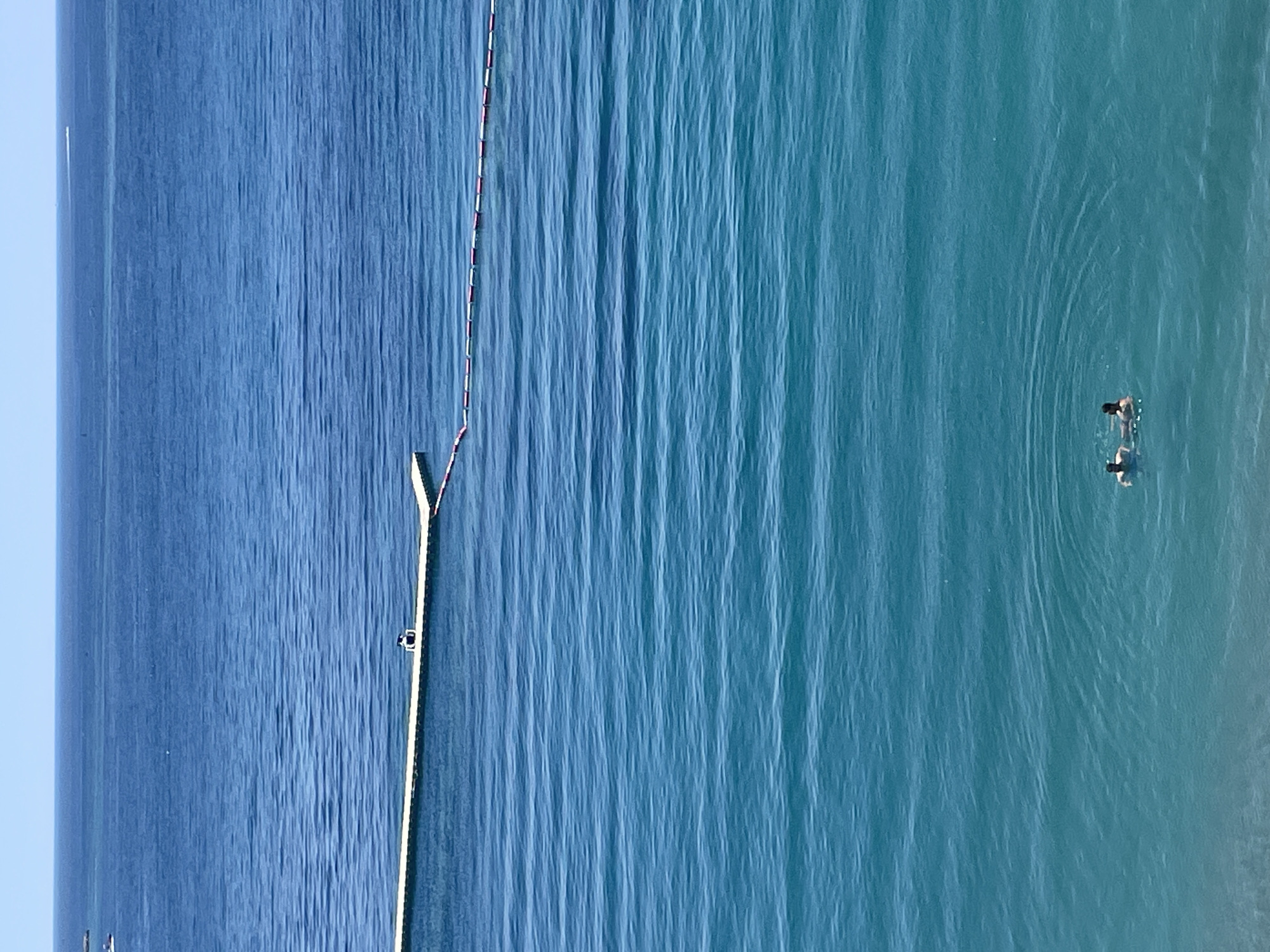 The image shows a body of water, likely the Adriatic Sea, near the city of Budva, Montenegro. A long, narrow barrier, possibly a floating dock or a safety line, extends into the water. Two people are swimming in the water away from the barrier. The water's color varies from dark blue to turquoise, suggesting different depths or light conditions. The sky is clear and bright.