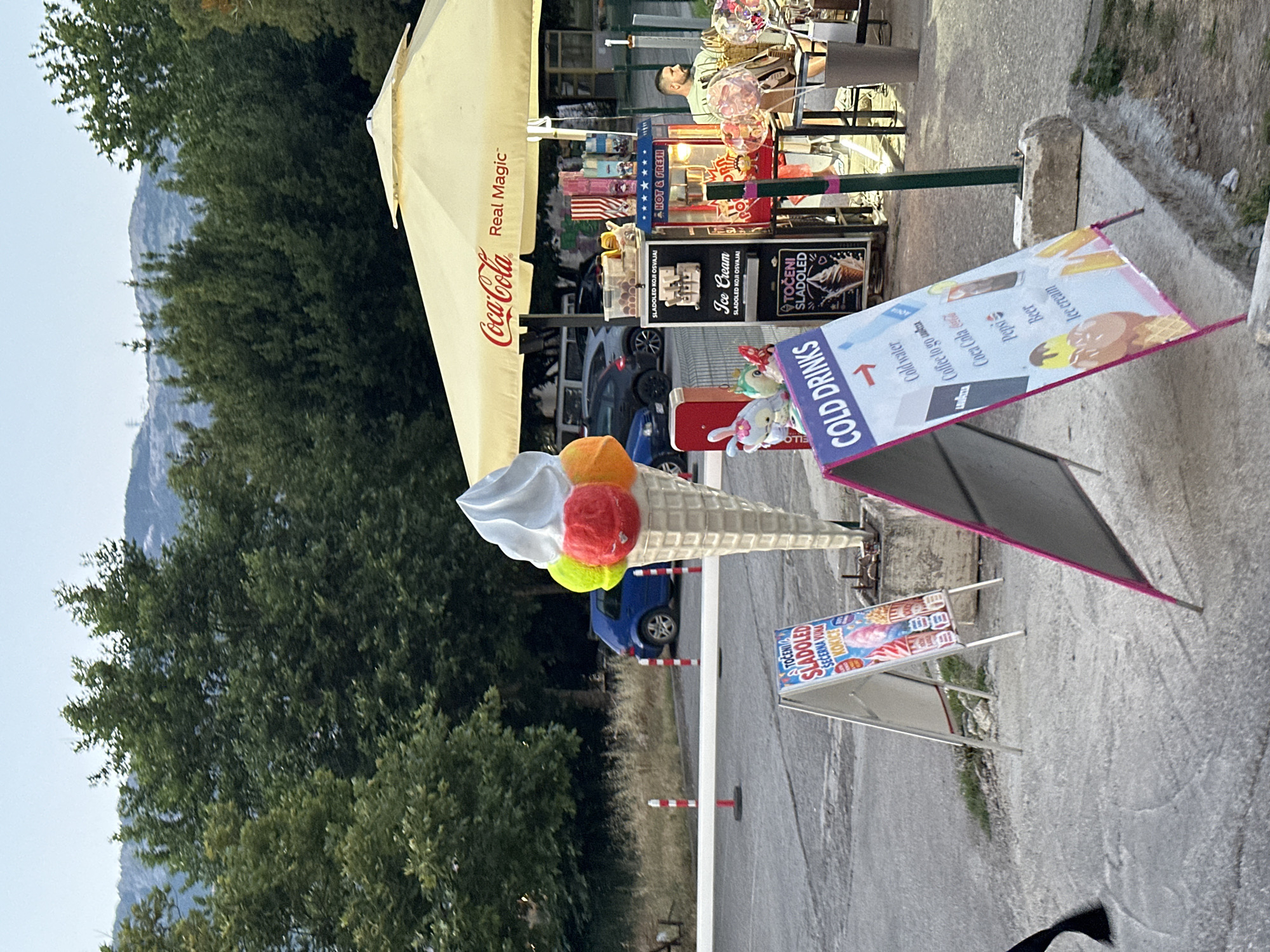 A street-level view captures an outdoor food stall in Budva, Montenegro, under a large yellow Coca-Cola branded umbrella. In the foreground, a pink-trimmed A-frame sign advertises "COLD DRINKS" with a list including cold water, coffee, Coca-Cola, Pepsi, beer, and ice cream, accompanied by illustrations of beverages and ice cream cones. To the left of this sign, another A-frame sign displays an advertisement for "SLADOLED SECERNA VUNA KOKICE", featuring images of ice cream and cotton candy. Dominating the center of the frame is a large, decorative ice cream cone sculpture with three scoops of ice cream in vibrant colors. Behind the sculpture, a blue car is parked in a lot, with trees and distant mountains forming the backdrop. The stall itself is equipped with a popcorn machine, an ice cream dispenser, and various display items. A man wearing a green shirt is seated at the stall, appearing to serve customers. The overall scene depicts a lively, open-air vendor area, likely a popular spot for snacks and refreshments.