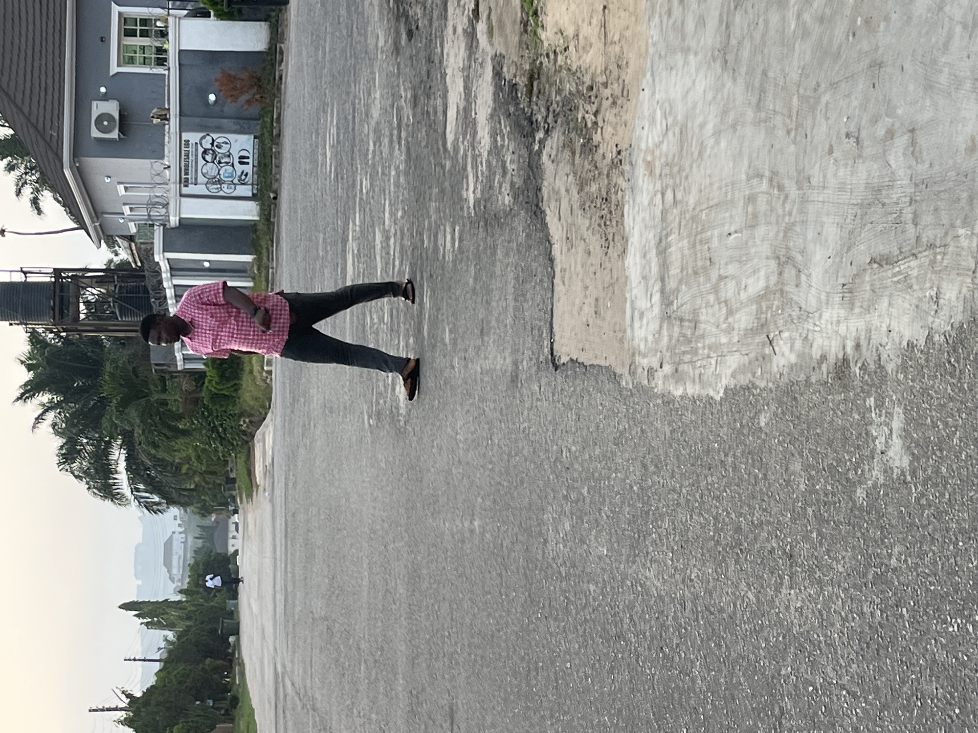 The image shows a man walking on a paved road in a residential area. The man is wearing a pink shirt and dark pants. A building with a sign is on the left. Palm trees are visible. In the distance, the city of Abuja, Nigeria is visible.