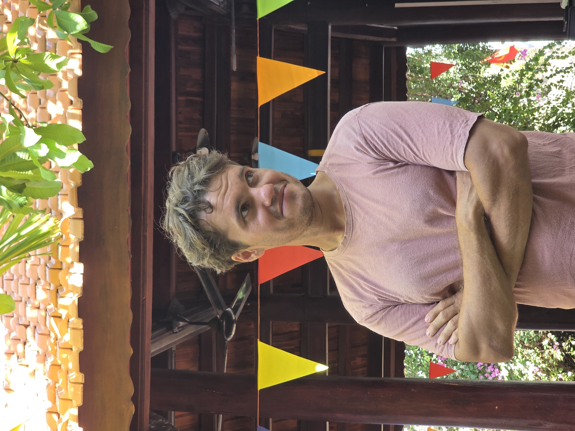 The image shows a man in a pink shirt with his arms crossed, standing in what appears to be an open-air structure decorated with colorful triangular flags. Behind him, through the openings of the structure, one can see green foliage and a hint of red flags. The architectural style suggests a tropical location. The man has curly hair that appears to be highlighted or lightened, and he is looking slightly up and to the left. The left side of the frame shows a tan lattice-like structure and green leaves. Based on the style of the structure and vegetation, this image is likely in Mũi Né, Vietnam.