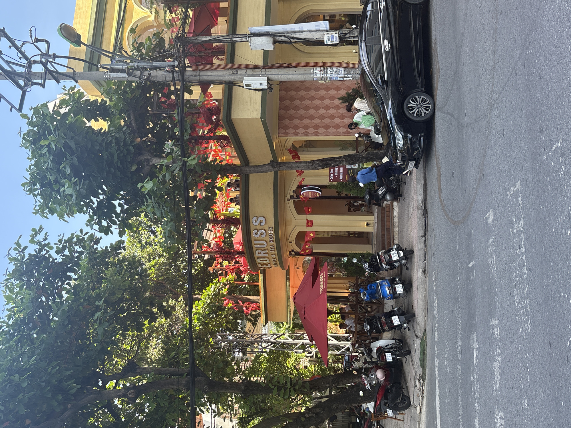 The image captures a street scene in Nha Trang Ward, Vietnam, featuring a brightly colored yellow building with "EDRUSS COFFEE & TEA HOUSE" signage prominently displayed. Red decorative flags and banners adorn the building's exterior, contributing to a festive atmosphere. In front of the establishment, several motorbikes are parked along the sidewalk. A black car is positioned in the foreground, facing right. Various individuals are visible, some seated at tables under a large red umbrella outside the coffee house, while others are standing near the building, interacting with each other. The environment includes lush green trees providing shade, overhead power lines, and streetlights. The sidewalk appears to be paved with concrete, and the street itself shows signs of wear. The sky is clear and blue, suggesting a sunny day.