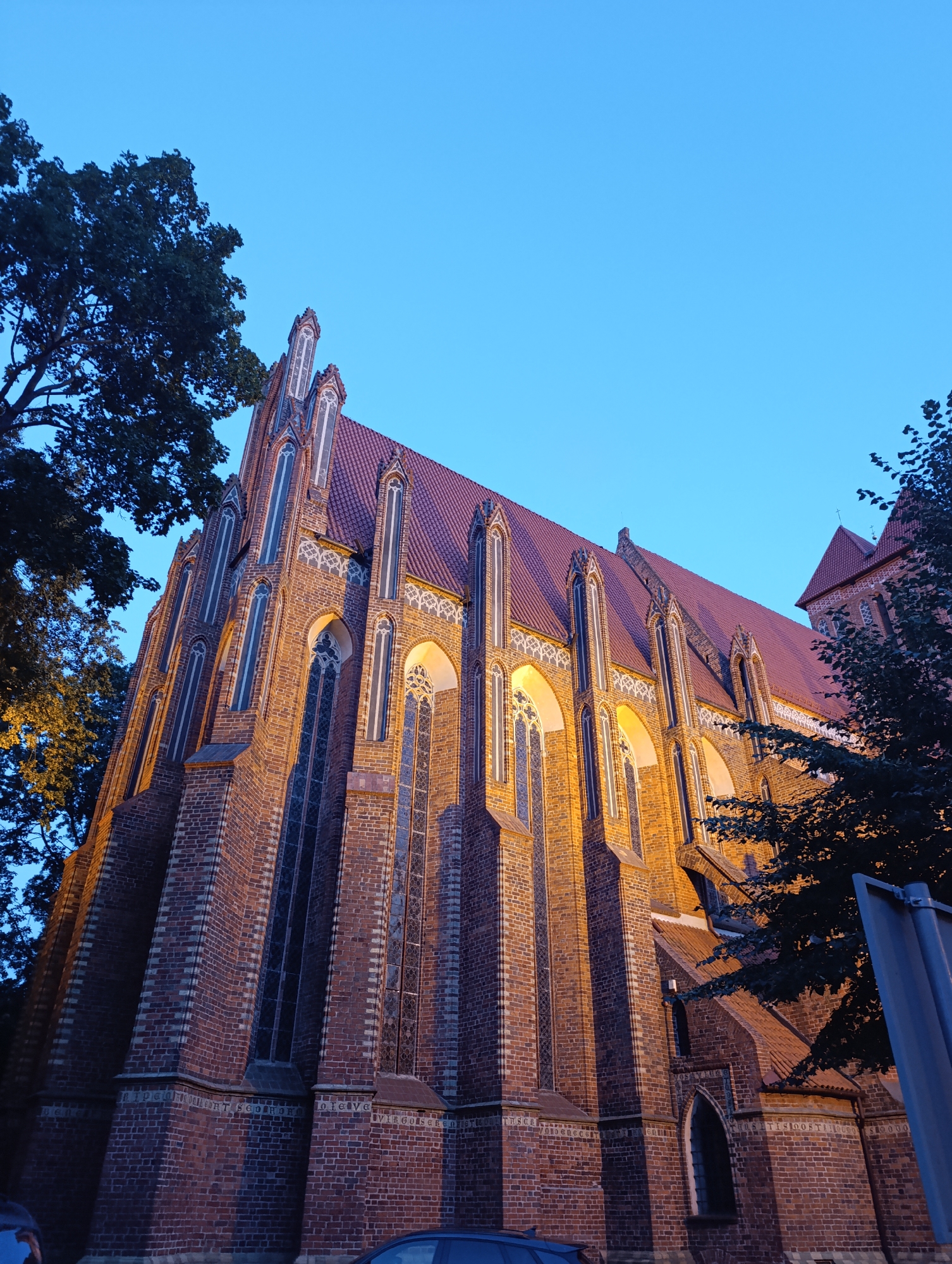 An exterior, low-angle, full-shot photograph of the Church of St. John the Baptist and St. John the Evangelist in Toruń, Poland, taken at dusk. The Gothic brick church is illuminated by warm artificial light. Tall, narrow arched windows with decorative tracery are visible along the facade. The red tiled roof slants upwards, and pointed spires top the buttresses. A large tree with dark green foliage is to the left of the church. The sky is a clear, deep blue, indicating twilight. To the right, a portion of another building, likely part of the same complex, is visible with a red pitched roof and a small tower topped with a cross. A dark colored car is parked in the lower-left corner of the image. A blue street sign is visible on the right side of the image.